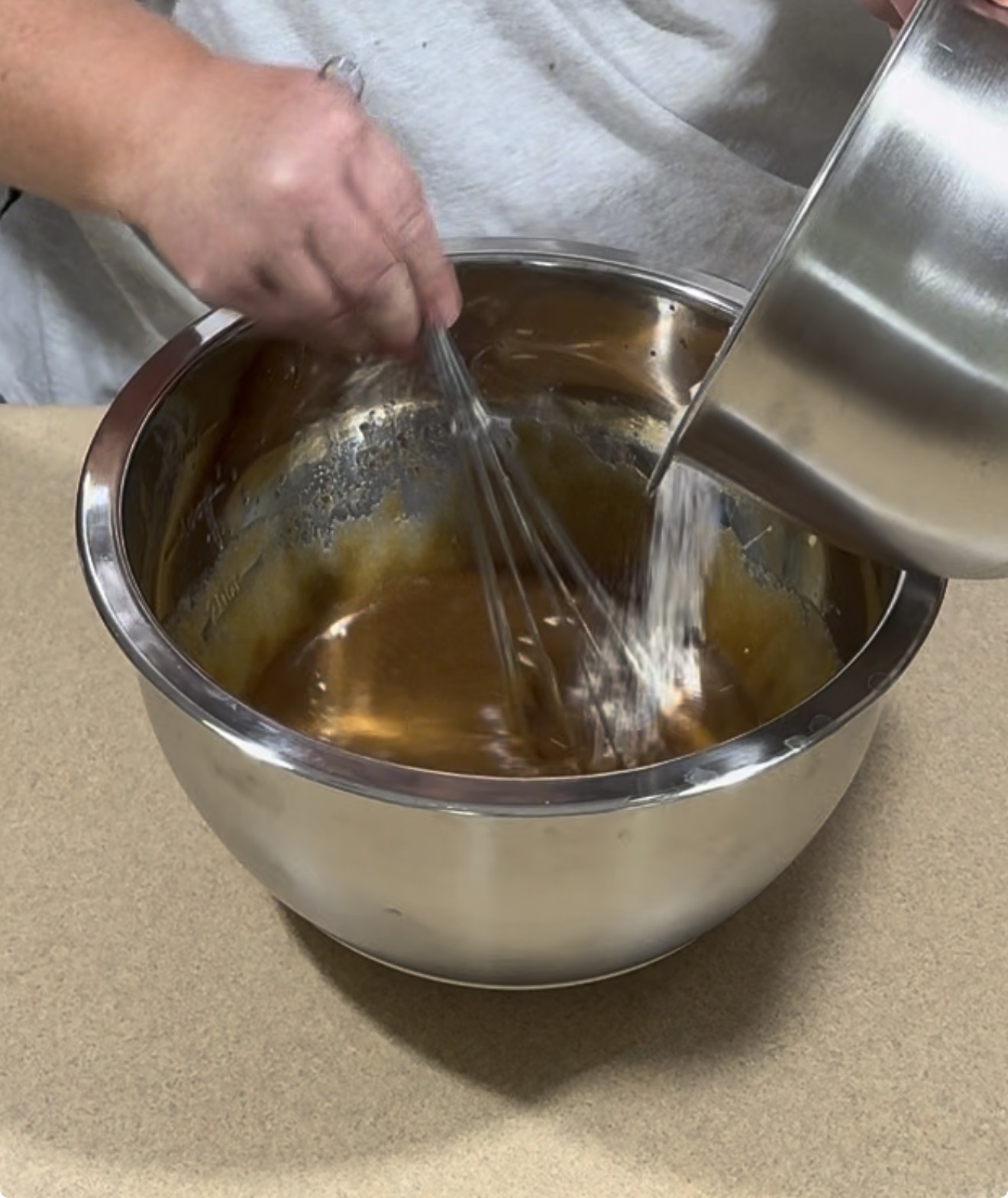 A person pours flour from a metal bowl into another bowl while whisking a brown mixture, preparing batter on a kitchen counter.