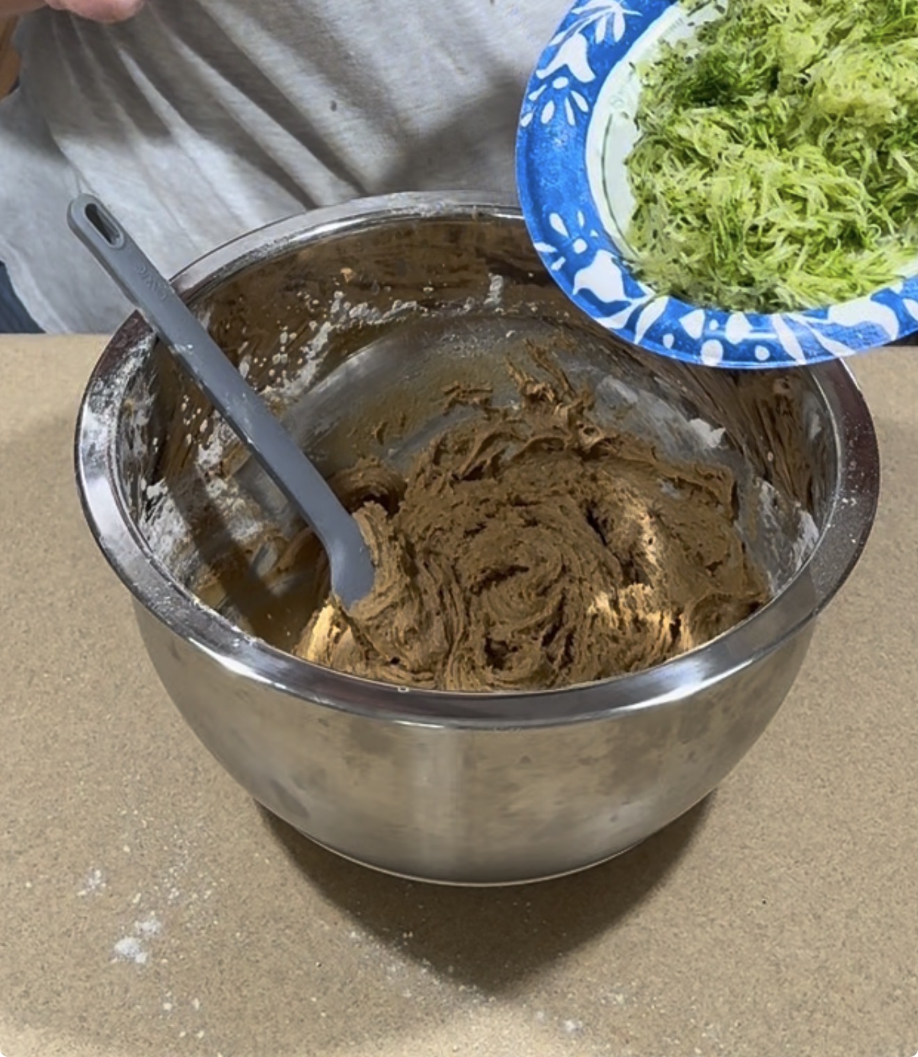 A person holds a plate of shredded zucchini over a mixing bowl filled with brown batter, preparing to add the zucchini. A gray spatula rests in the batter.