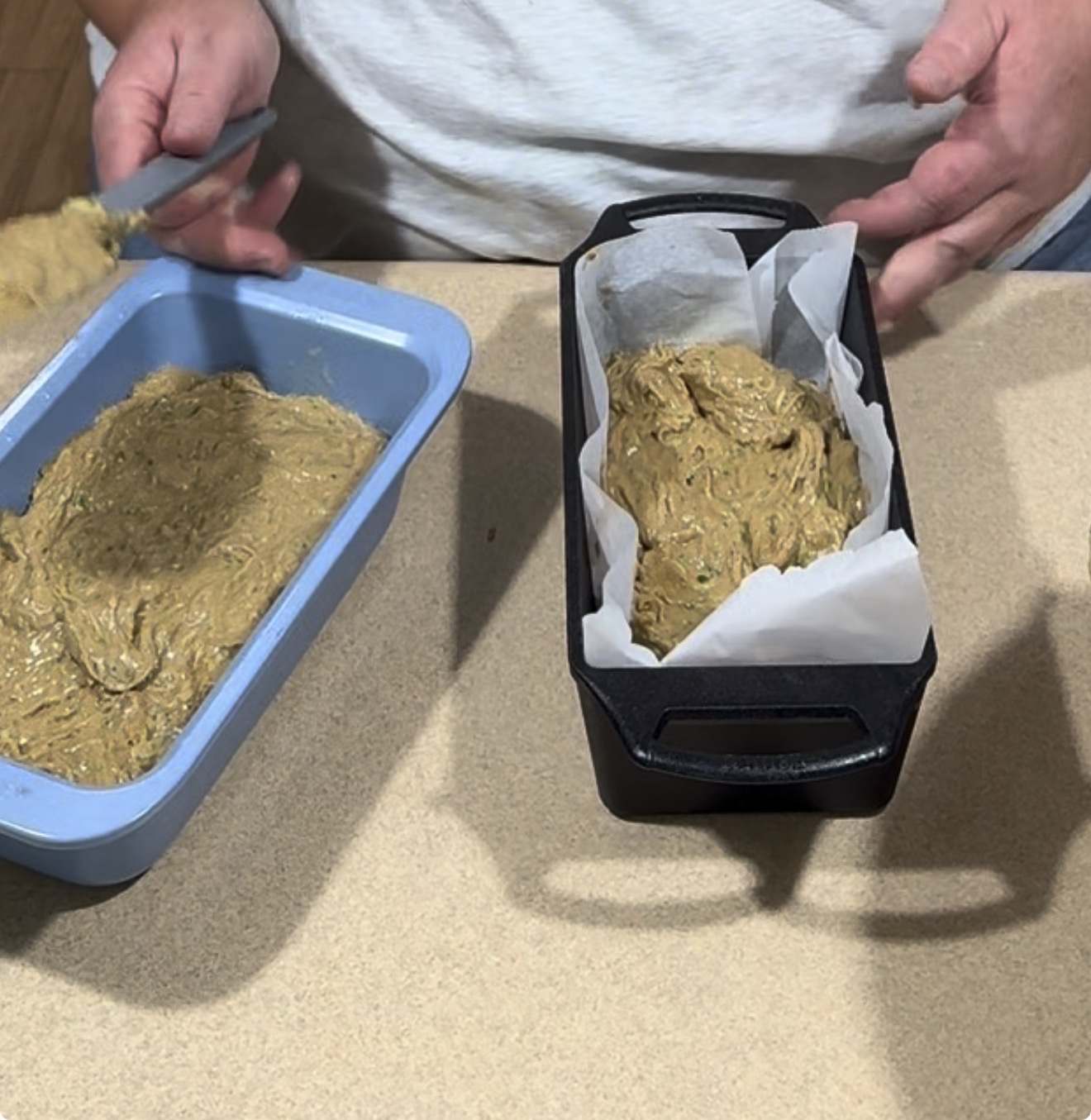 Two loaf pans on a counter, one blue and one black with parchment paper, being filled with raw batter by a person wearing a white shirt.