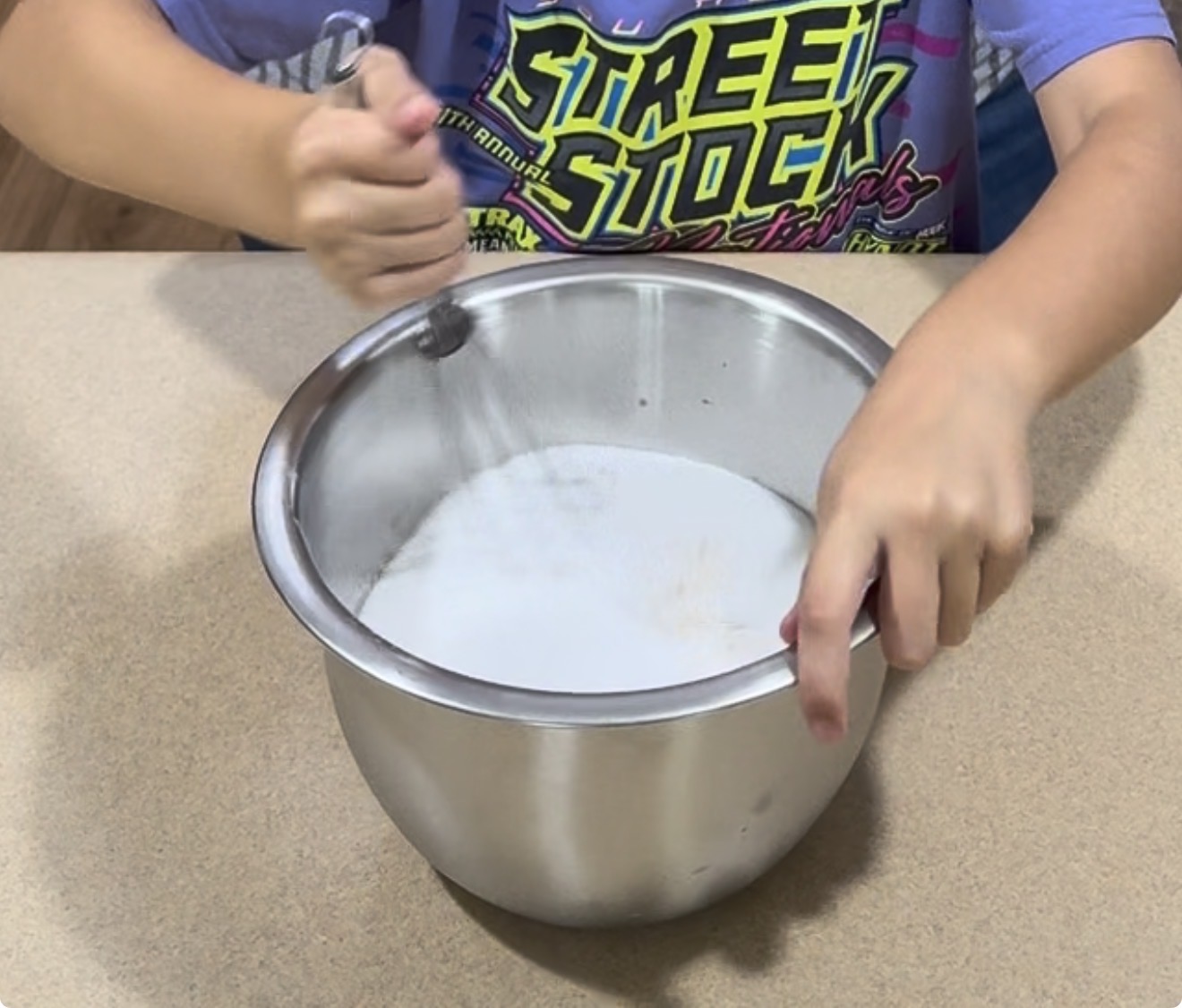 A person stirs white sugar in a large metal bowl with a whisk, wearing a colorful Street Stock T-shirt, on a light brown countertop.