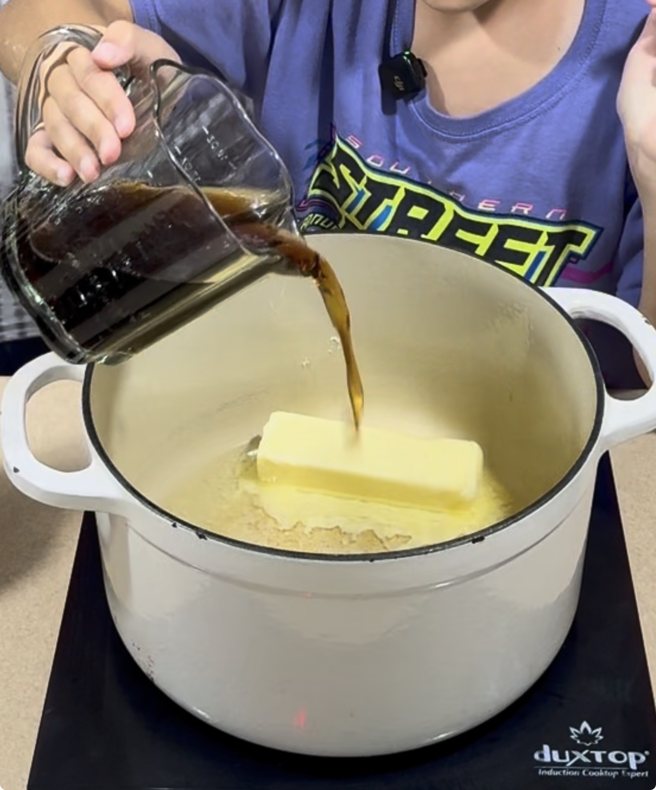 A person pours a dark liquid from a glass measuring cup into a white pot with a stick of butter on a stovetop.