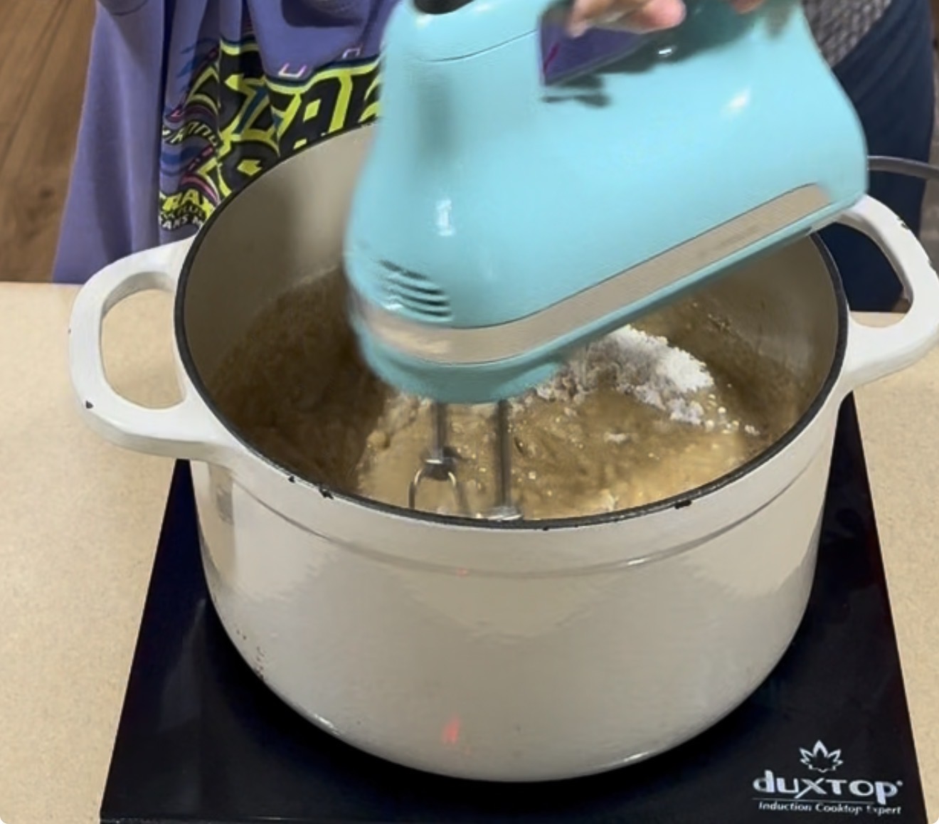 A person uses a blue electric hand mixer to blend ingredients in a large white pot on a black induction cooktop.