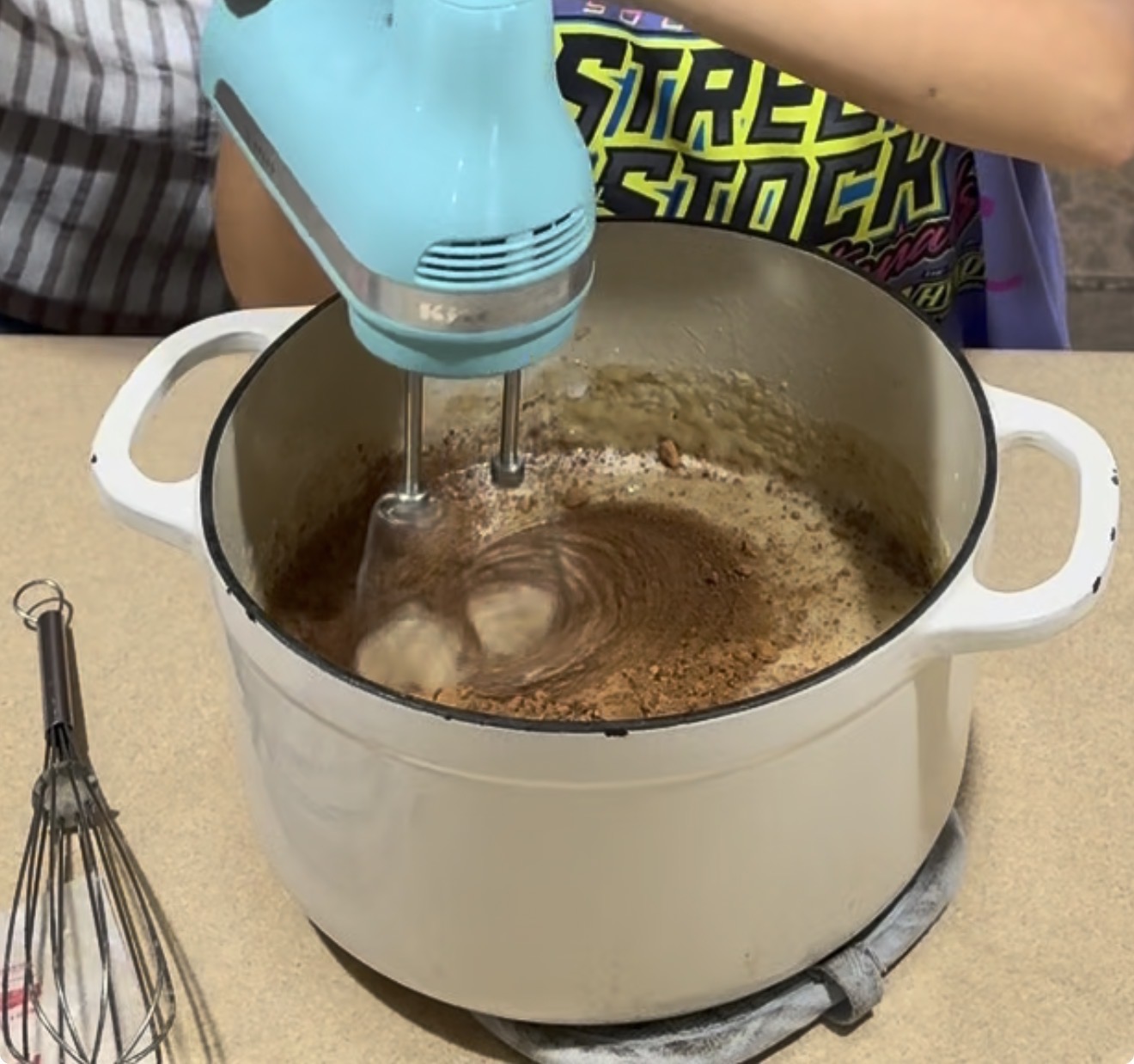 A person uses a blue hand mixer to blend cocoa powder in a white pot on a stovetop, with a metal whisk resting nearby.