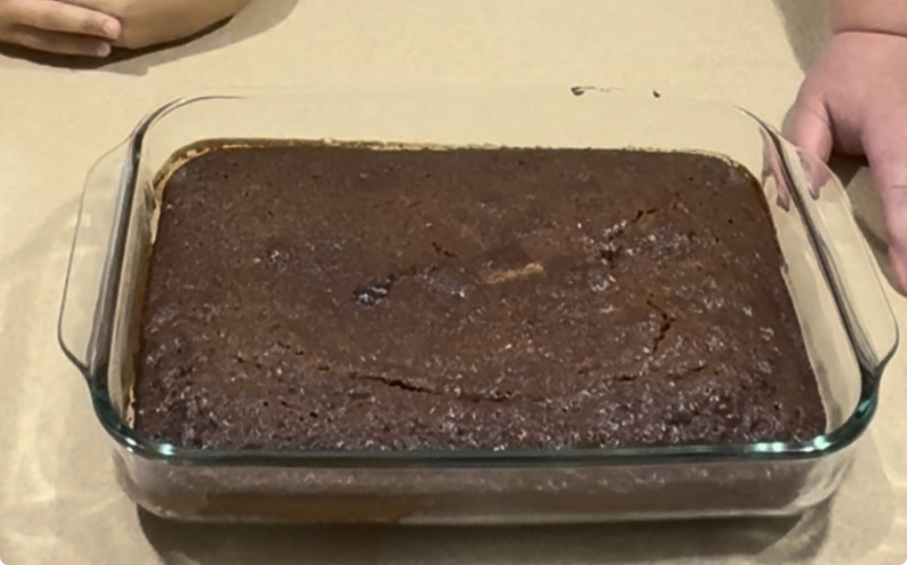 A rectangular glass baking dish holds a freshly baked chocolate cake, with two hands resting near the sides on a beige countertop.
