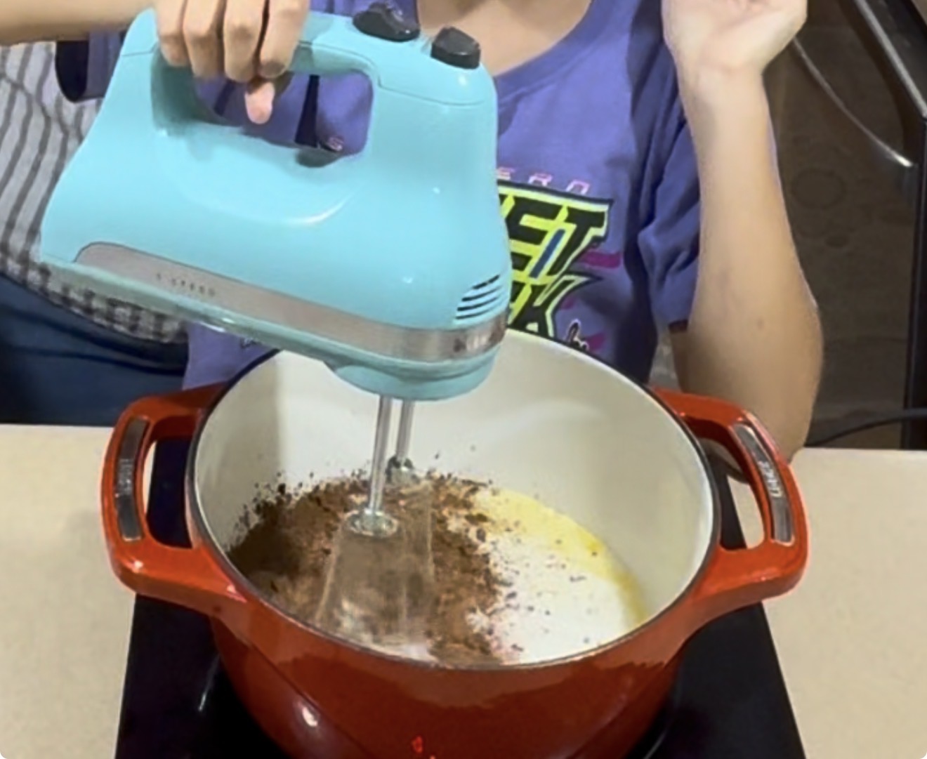 A person uses a blue electric hand mixer to blend ingredients, including cocoa powder, in a red pot on a stovetop.