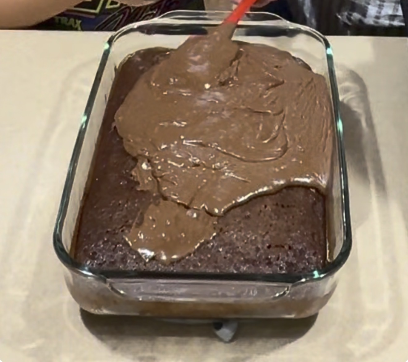 A glass baking dish with a rectangular chocolate cake. A person is spreading chocolate frosting on top with a red spatula. The dish is on a beige surface.