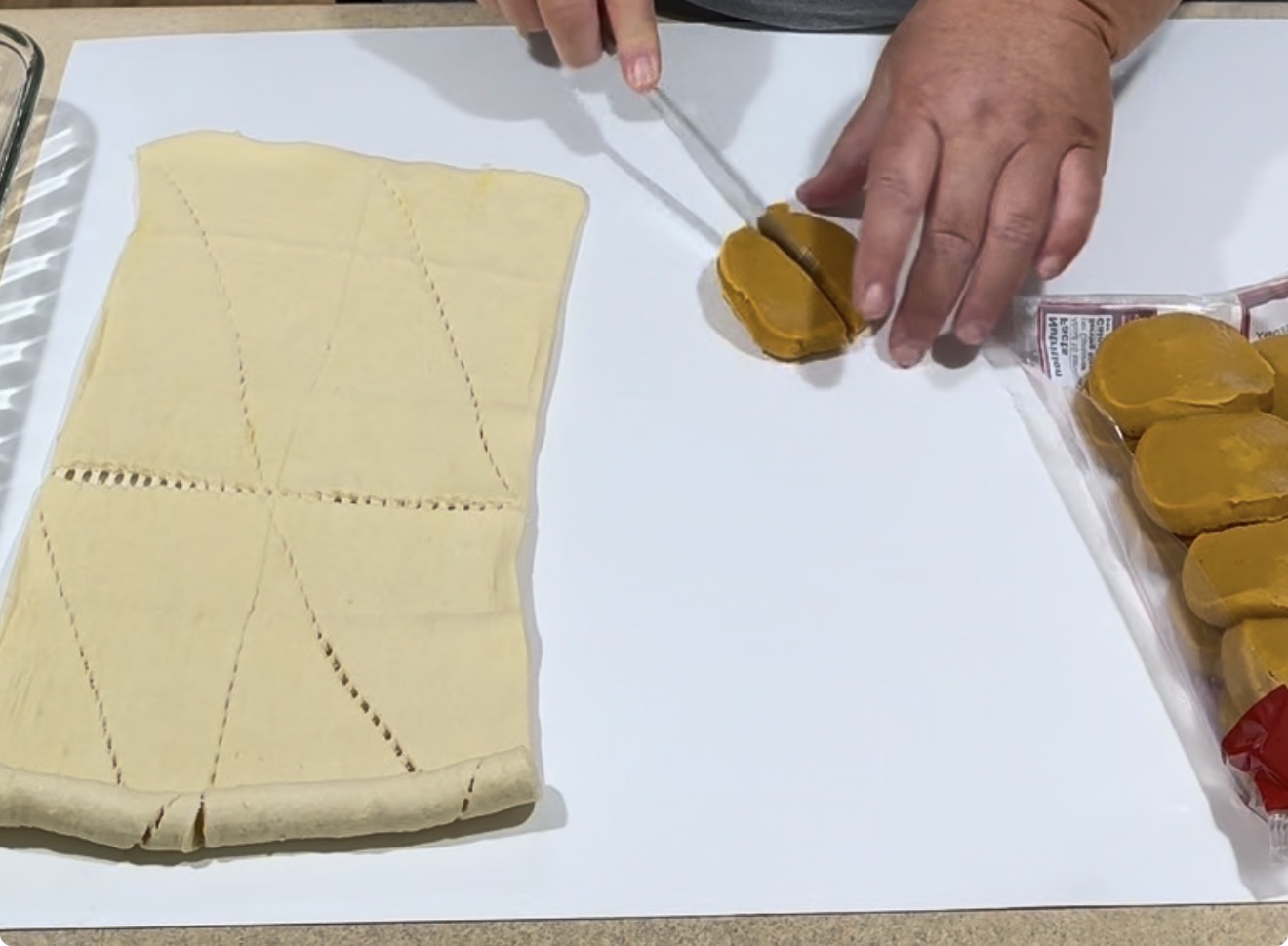 Hands slicing pumpkin spice cookie dough; next to it is a rectangle of perforated crescent roll dough on a white surface. The cookie dough package is partially visible on the right.