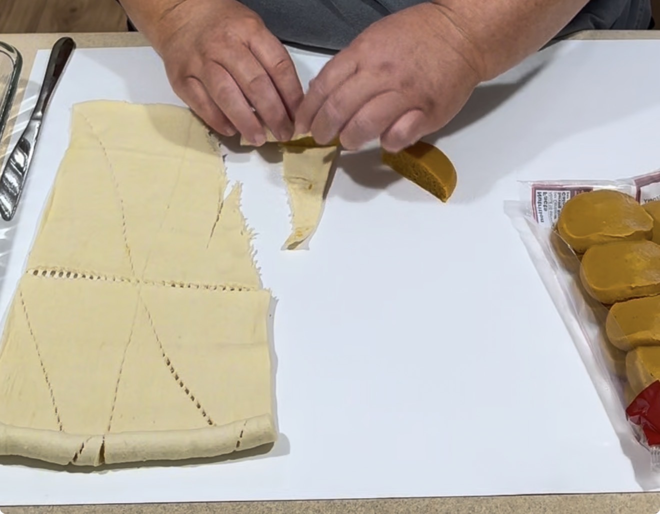 Hands rolling candied yams in crescent roll dough on a white surface, with a package of yams and utensils nearby. The dough is partially separated into triangles for wrapping.