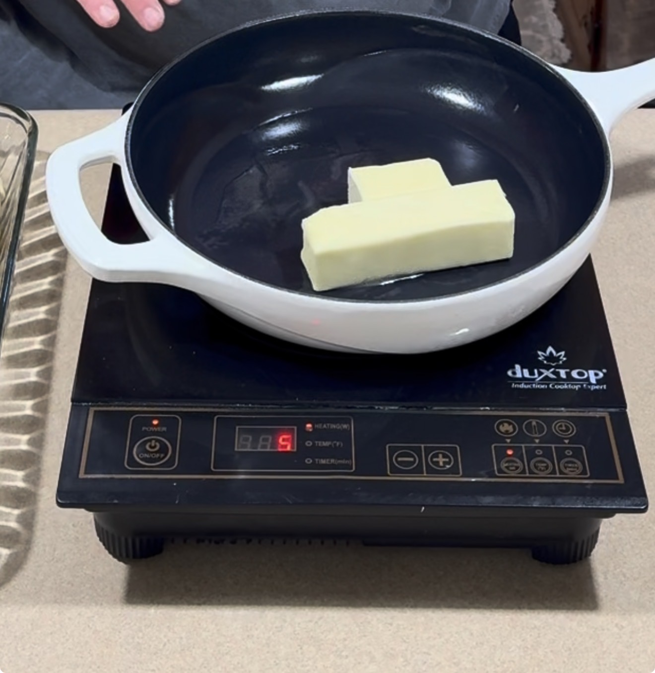 A white skillet with two sticks of butter sits on an induction cooktop set to level 5. A glass baking dish and a persons hands are visible in the background.