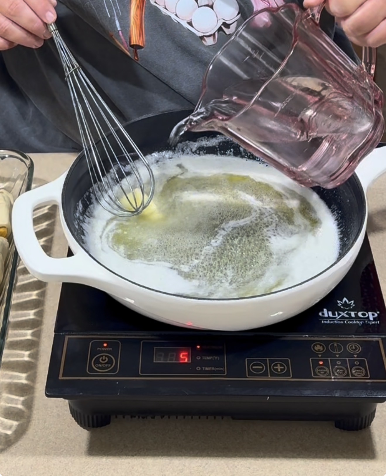 A person pours liquid from a measuring cup into a white pot with melted butter, stirring with a whisk on an induction cooktop.
