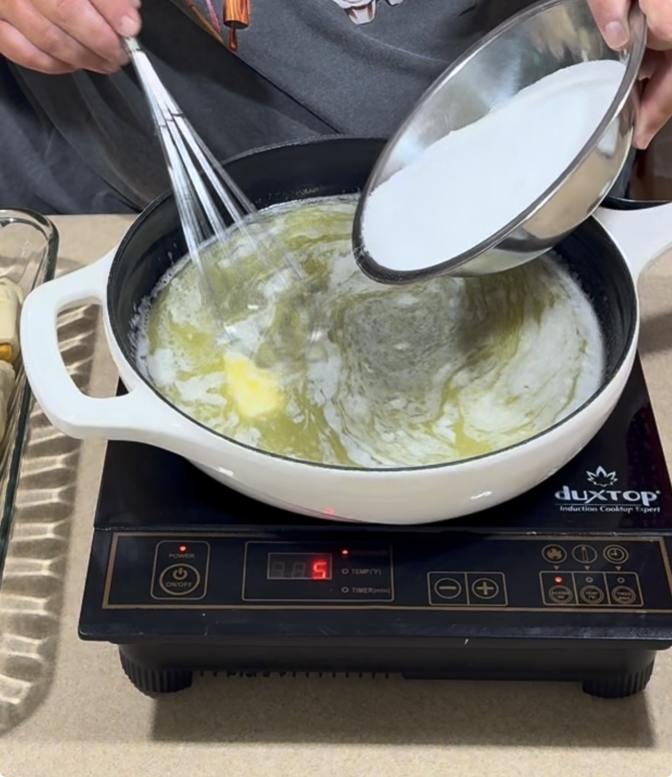 A person pours sugar into a white pot of melted butter on an induction cooktop, whisking the mixture. The cook wears a gray shirt, and a glass dish sits beside the stove.