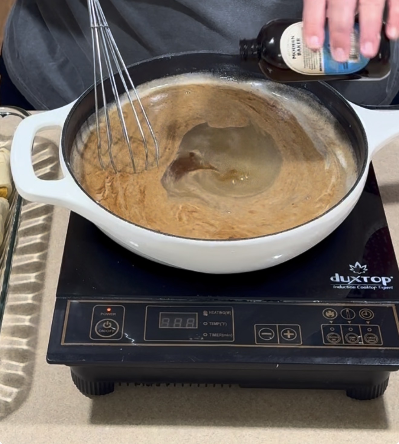 A person pours vanilla extract into a white pot of brown bubbling liquid on a portable induction cooktop, while whisking the mixture.
