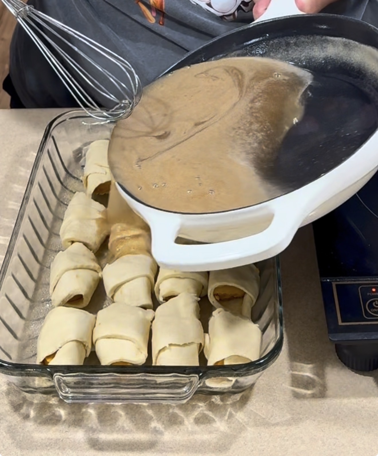 A person pours a creamy sauce from a white pan over crescent roll-wrapped pieces arranged in a glass baking dish on a kitchen counter.