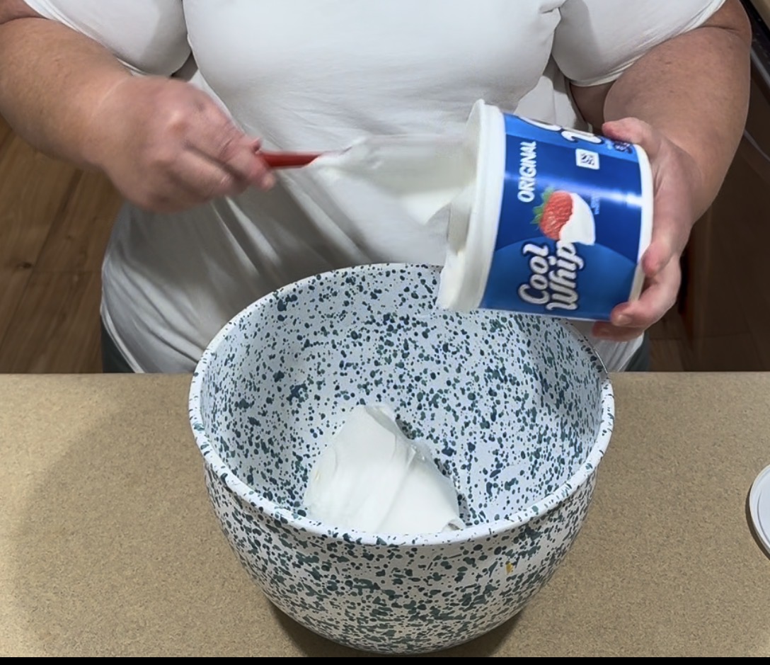 A person scoops Cool Whip from its container into a speckled mixing bowl on a kitchen counter.