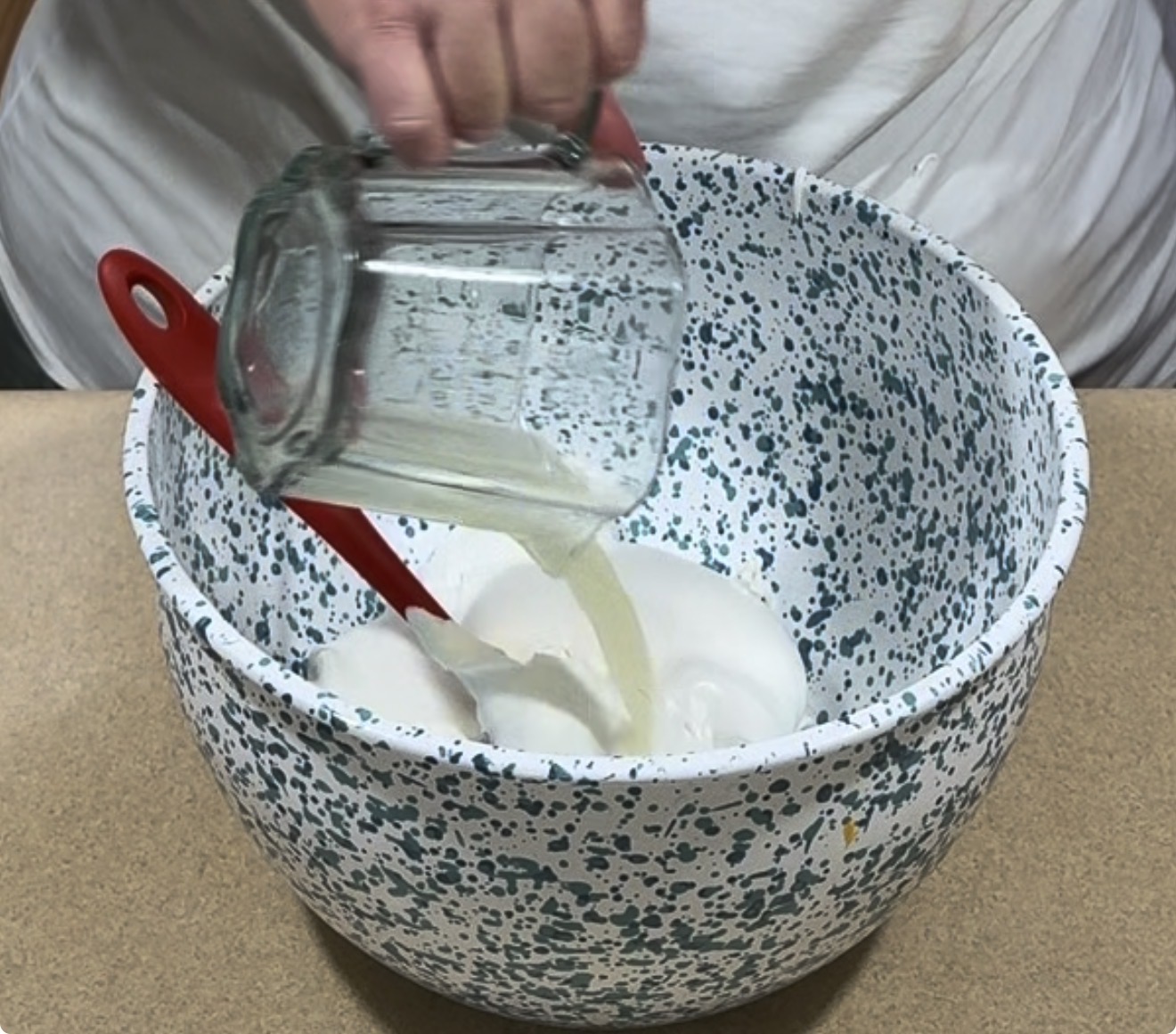 A person pours liquid from a glass measuring cup into a blue-speckled mixing bowl with a red spatula and white mixture inside, on a countertop.
