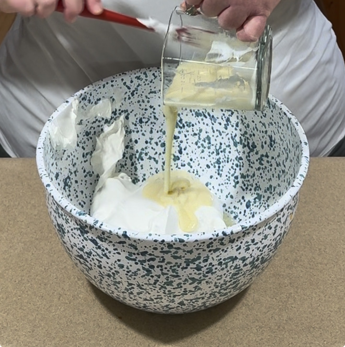 A person pours a thick yellow liquid from a glass into a speckled mixing bowl filled with a white, fluffy mixture, holding a red spatula in the other hand.