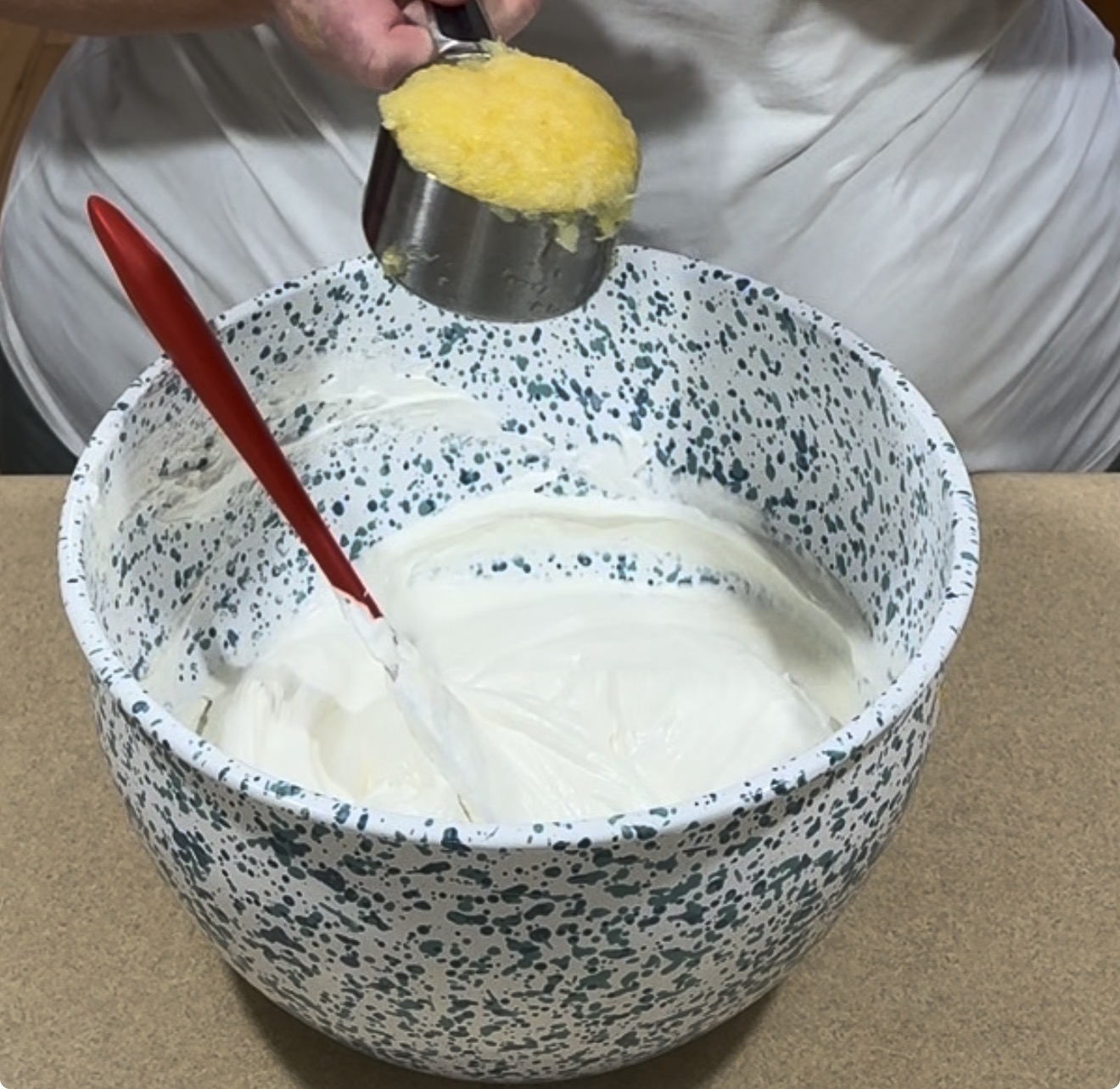 A person holds a measuring cup of yellow mixture above a large speckled bowl filled with white batter. A red spatula rests inside the bowl on a countertop.