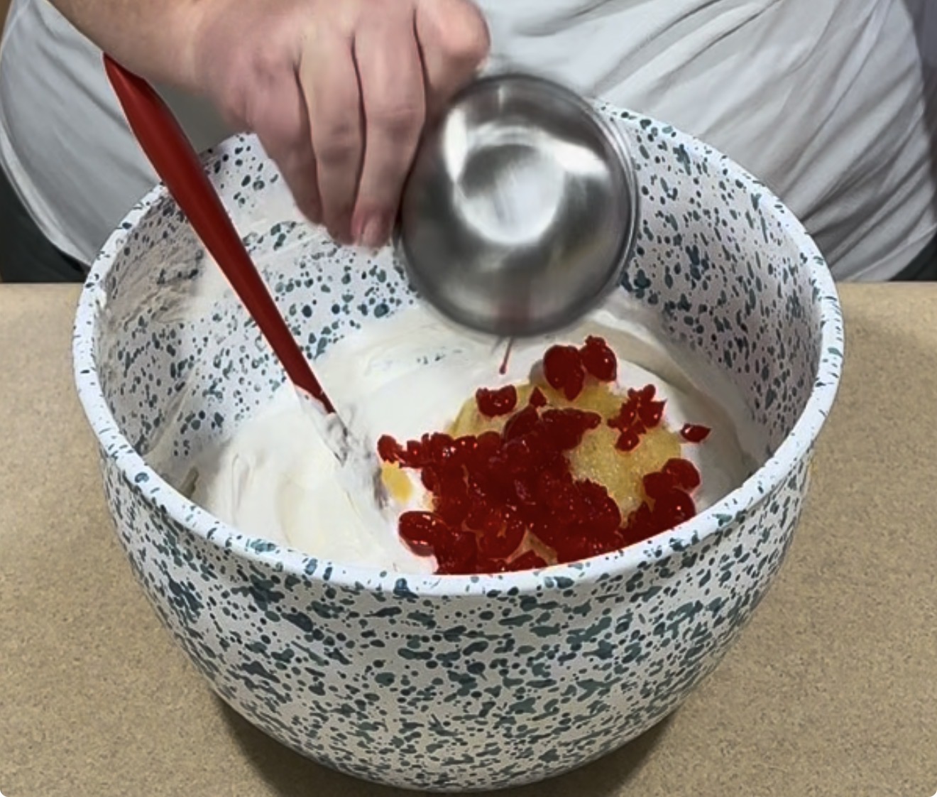 A person pours chopped red cherries from a small metal cup into a large speckled mixing bowl filled with white whipped mixture, using a red spatula to stir.