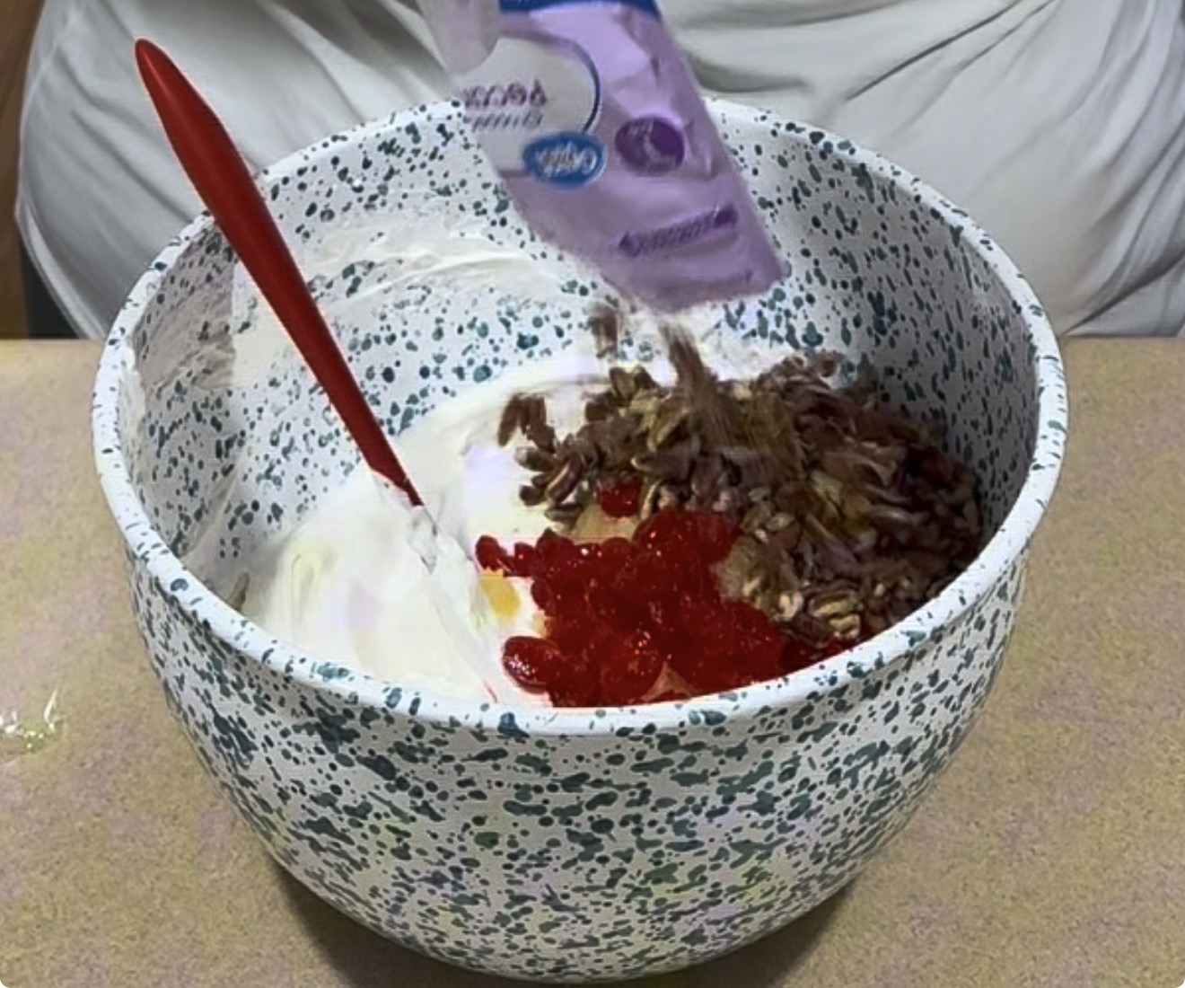 A person pours chopped nuts and other ingredients into a speckled mixing bowl filled with whipped cream. A red spatula rests inside the bowl, and bright red cherries are visible among the ingredients.