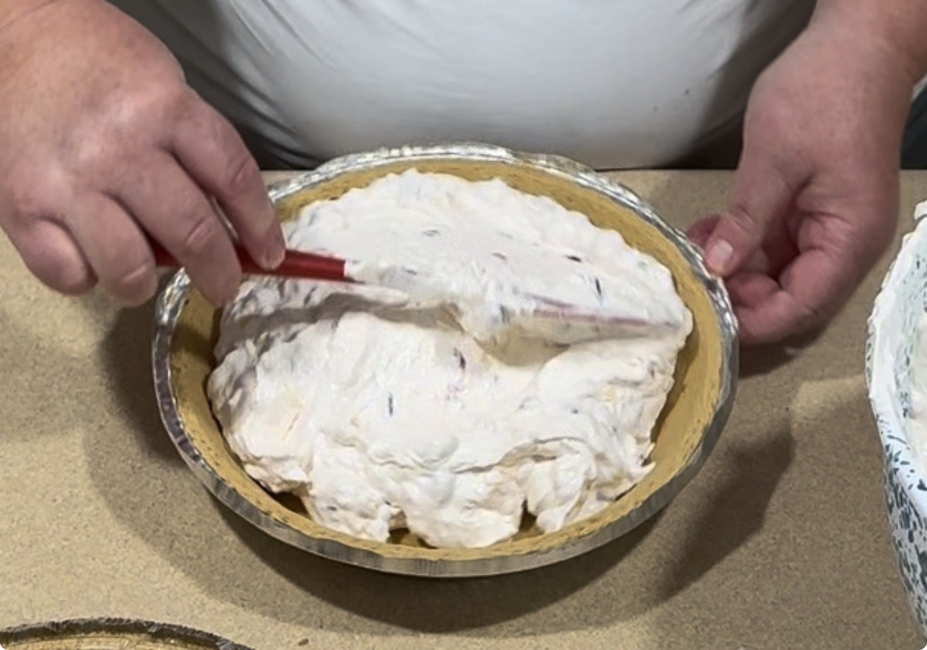 A person spreads a creamy pie filling with a spatula into a pie crust on a countertop.