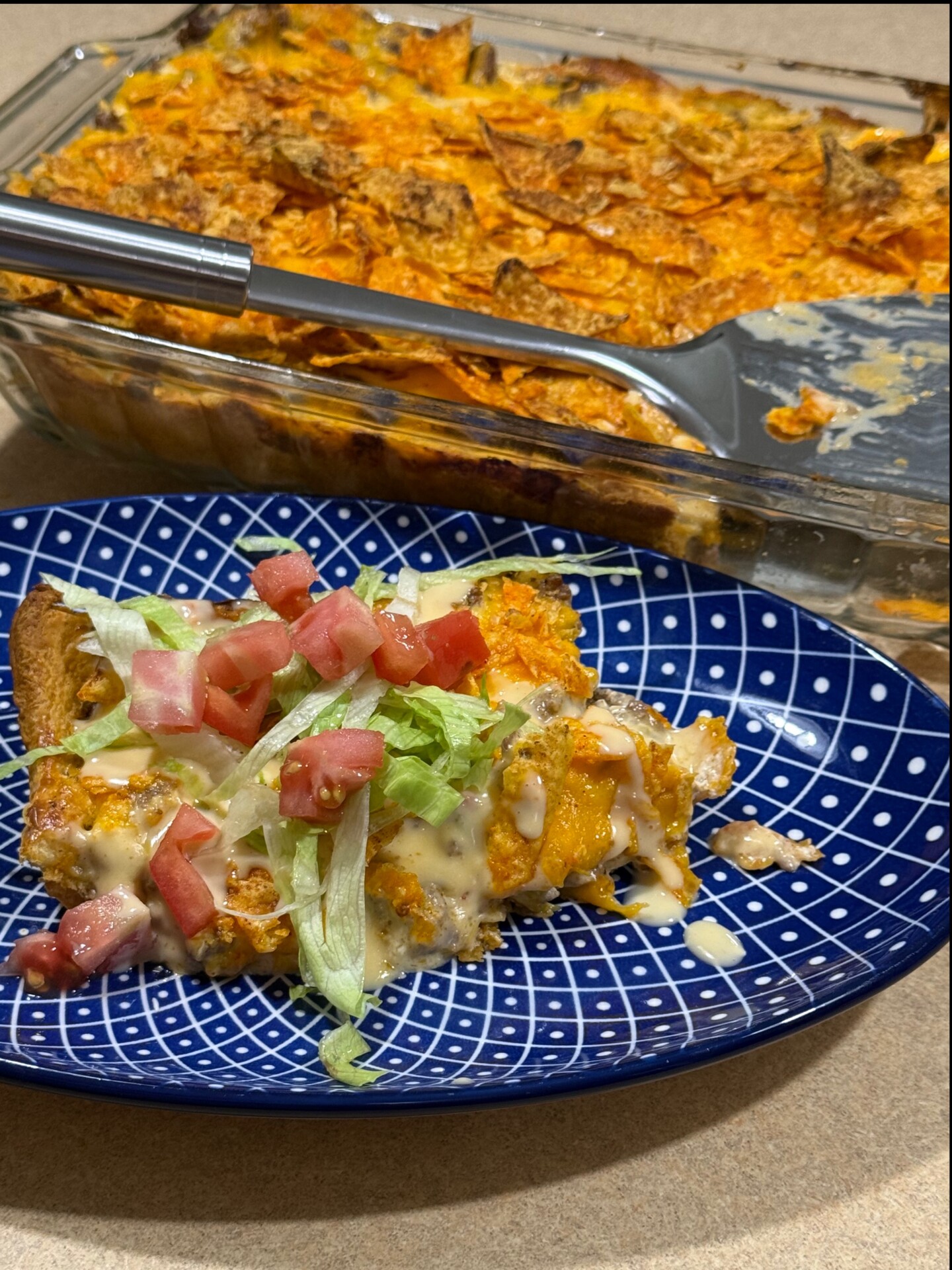 A slice of cheesy casserole with lettuce, tomato, and sauce sits on a blue patterned plate. The rest of the casserole is in a glass baking dish behind it with a serving spatula.