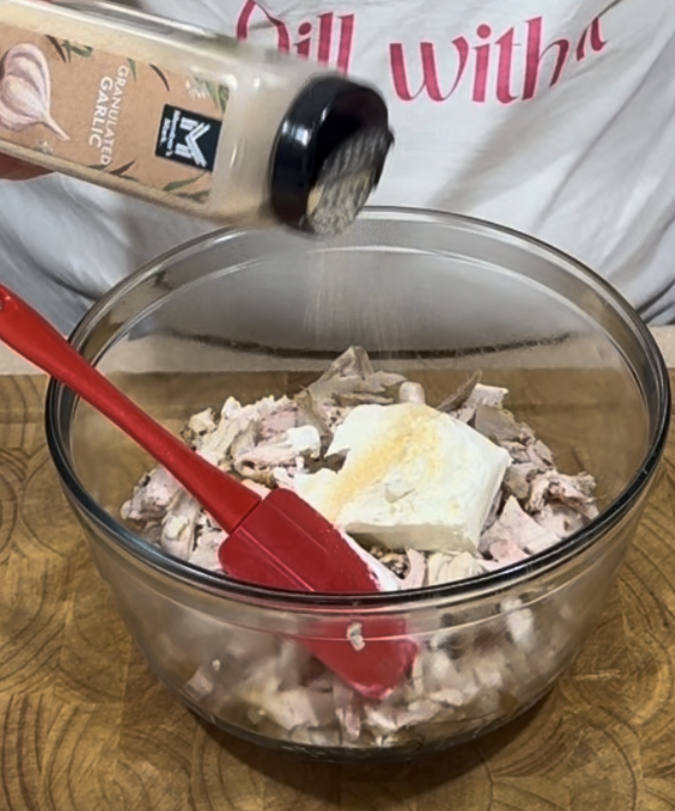 A person sprinkles garlic powder into a glass bowl containing chopped chicken and a block of cream cheese, using a red spatula to mix.