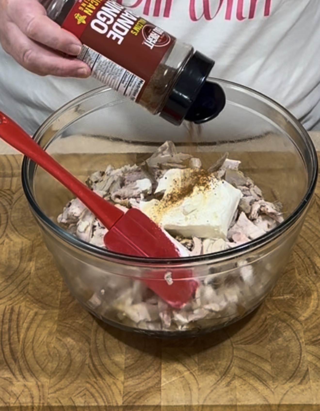 A person sprinkles seasoning from a bottle into a glass bowl filled with chopped meat and a block of cream cheese, mixing with a red spatula on a wooden surface.