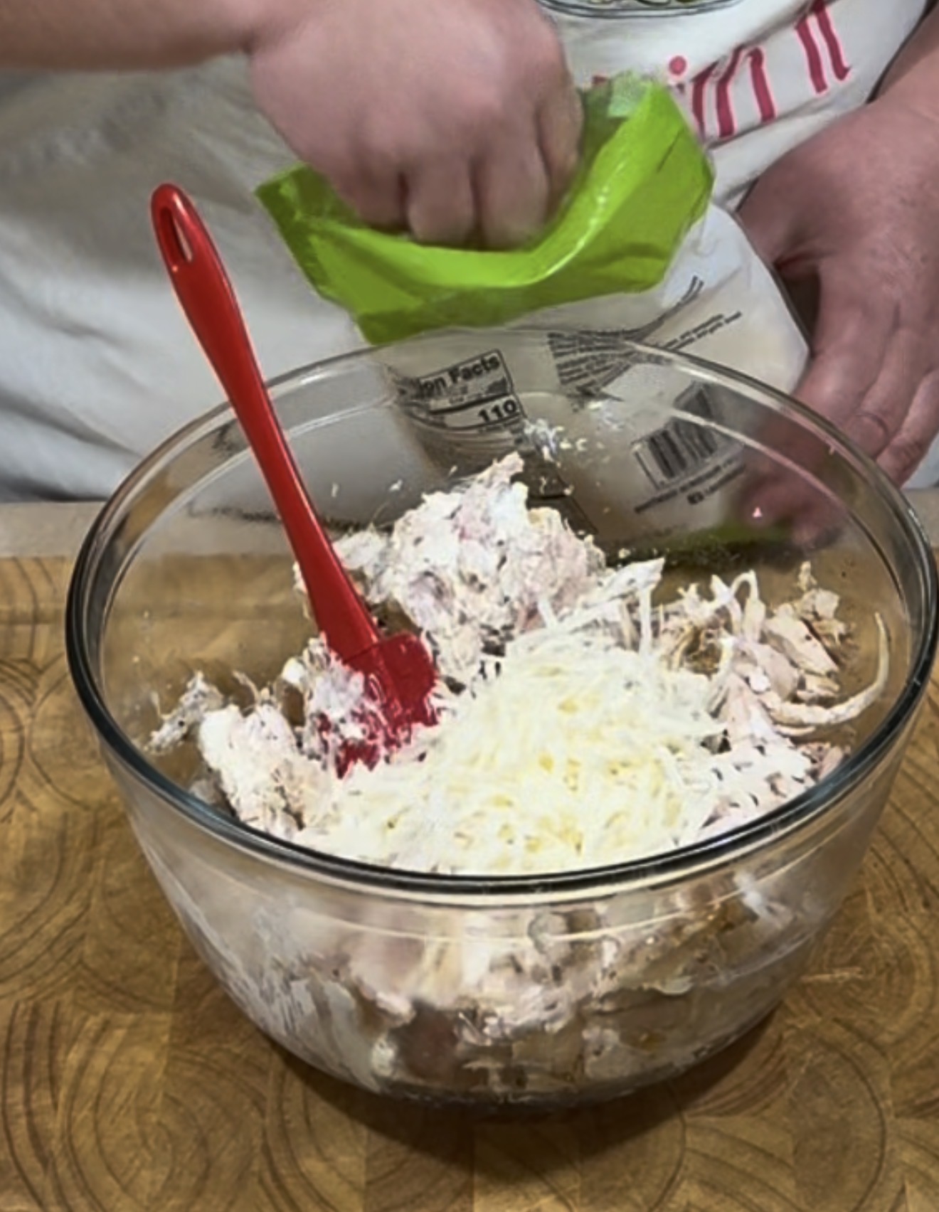 A person adds shredded cheese to a glass bowl filled with a creamy mixture, using a red spatula to stir, on a wooden surface.