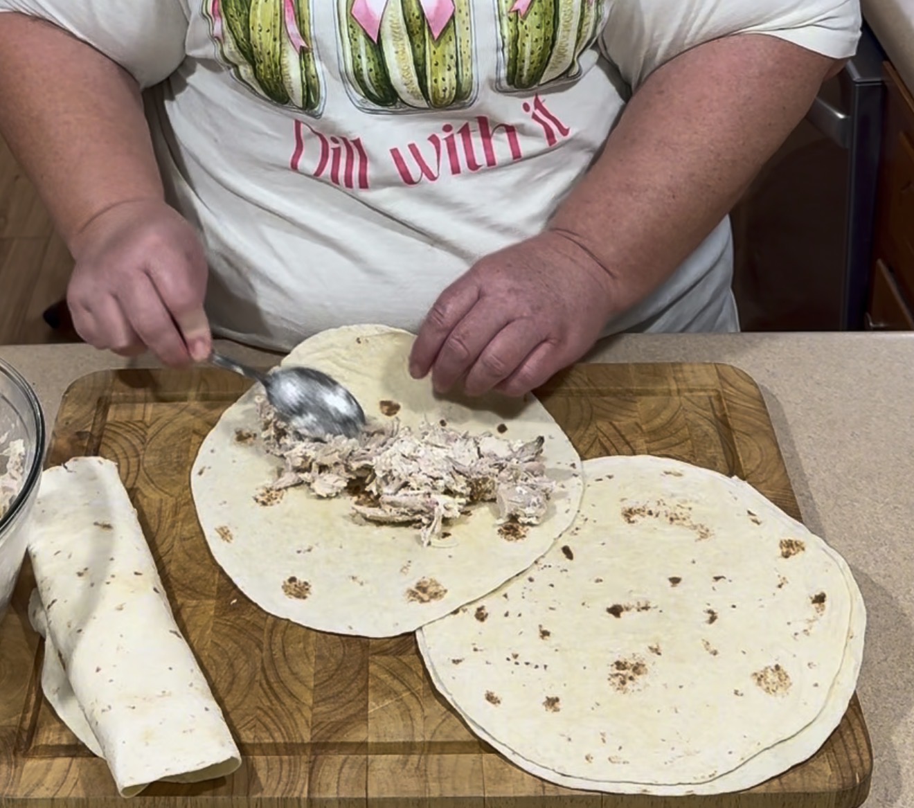 A person in a Dill with it shirt spoons shredded chicken onto a tortilla on a wooden board, with another tortilla and a rolled wrap beside it.