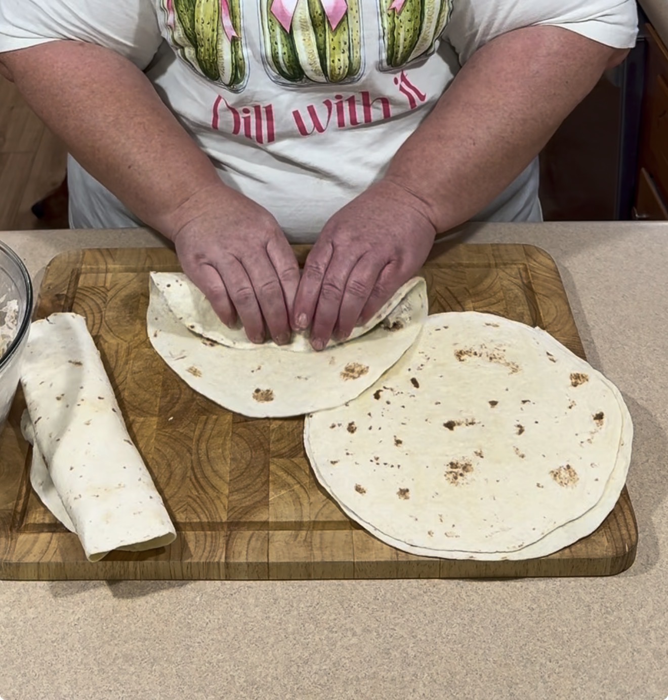 A person rolls a flour tortilla with filling on a wooden cutting board, next to another rolled tortilla and an unrolled tortilla. Both hands are visible, and the person wears a t-shirt with cacti illustrations.