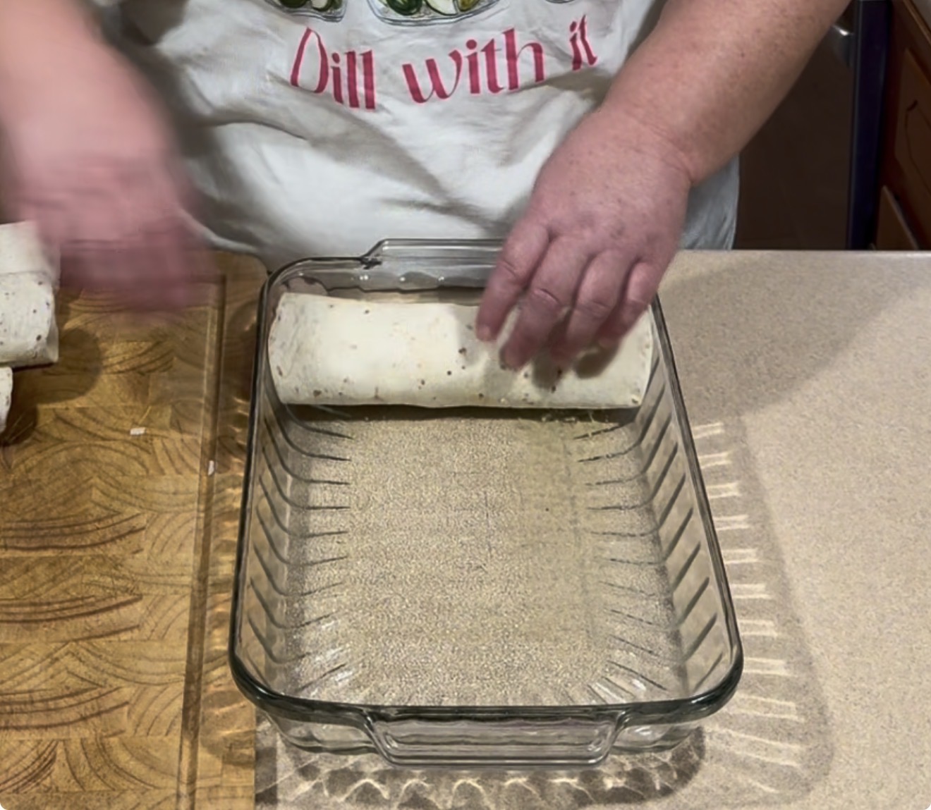 A person wearing a Dill with it apron places a rolled tortilla into a glass baking dish on a kitchen counter next to a wooden cutting board.