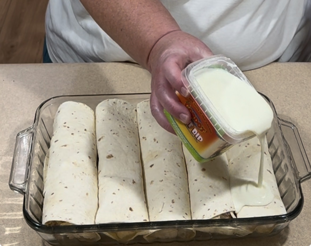 A person pours white cheese dip from a container onto rolled tortillas arranged in a glass baking dish on a kitchen counter.