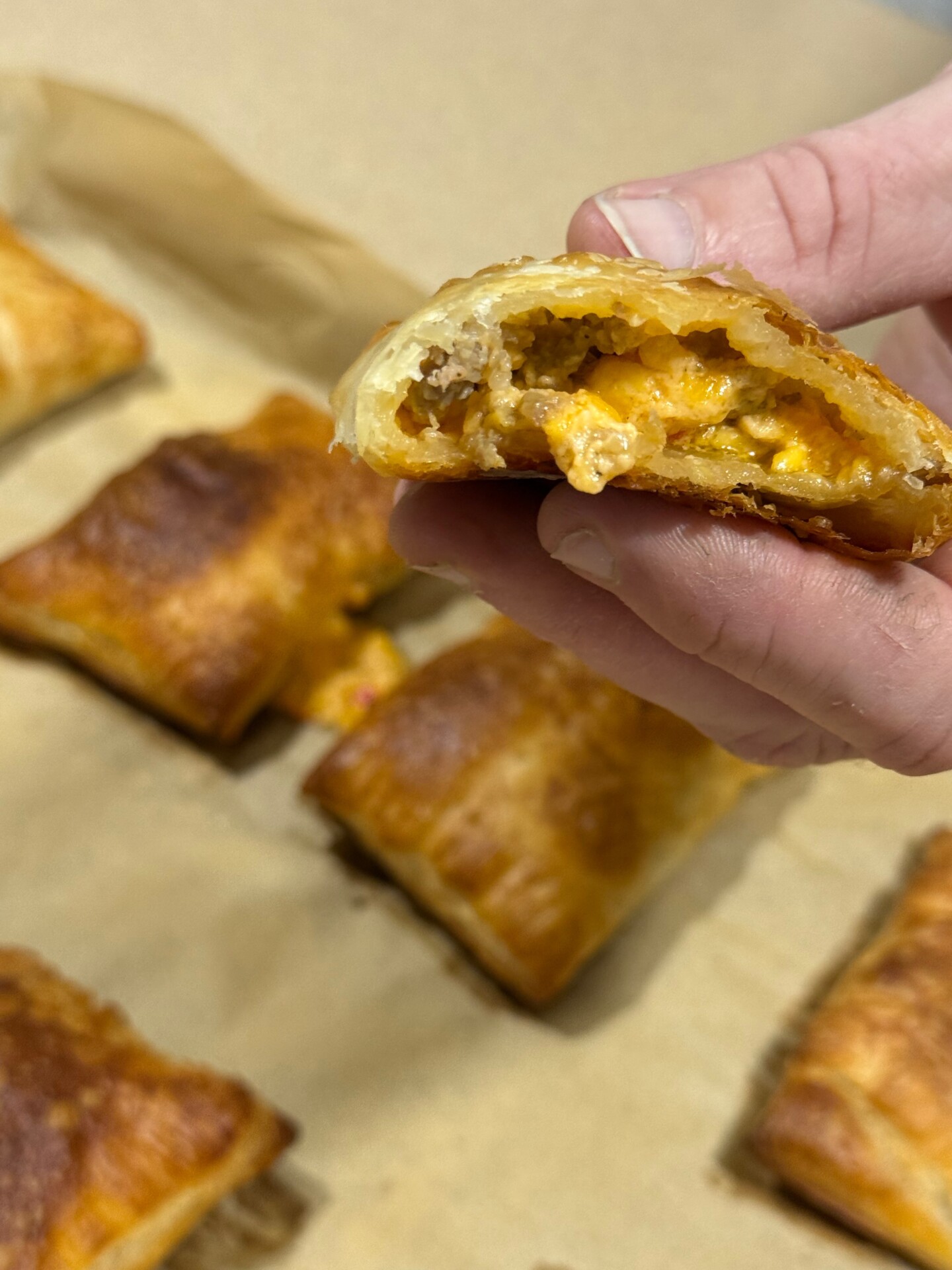 A hand holds a pastry with a flaky golden crust and a cheesy filling. More similar pastries rest on a parchment-lined baking sheet in the background.