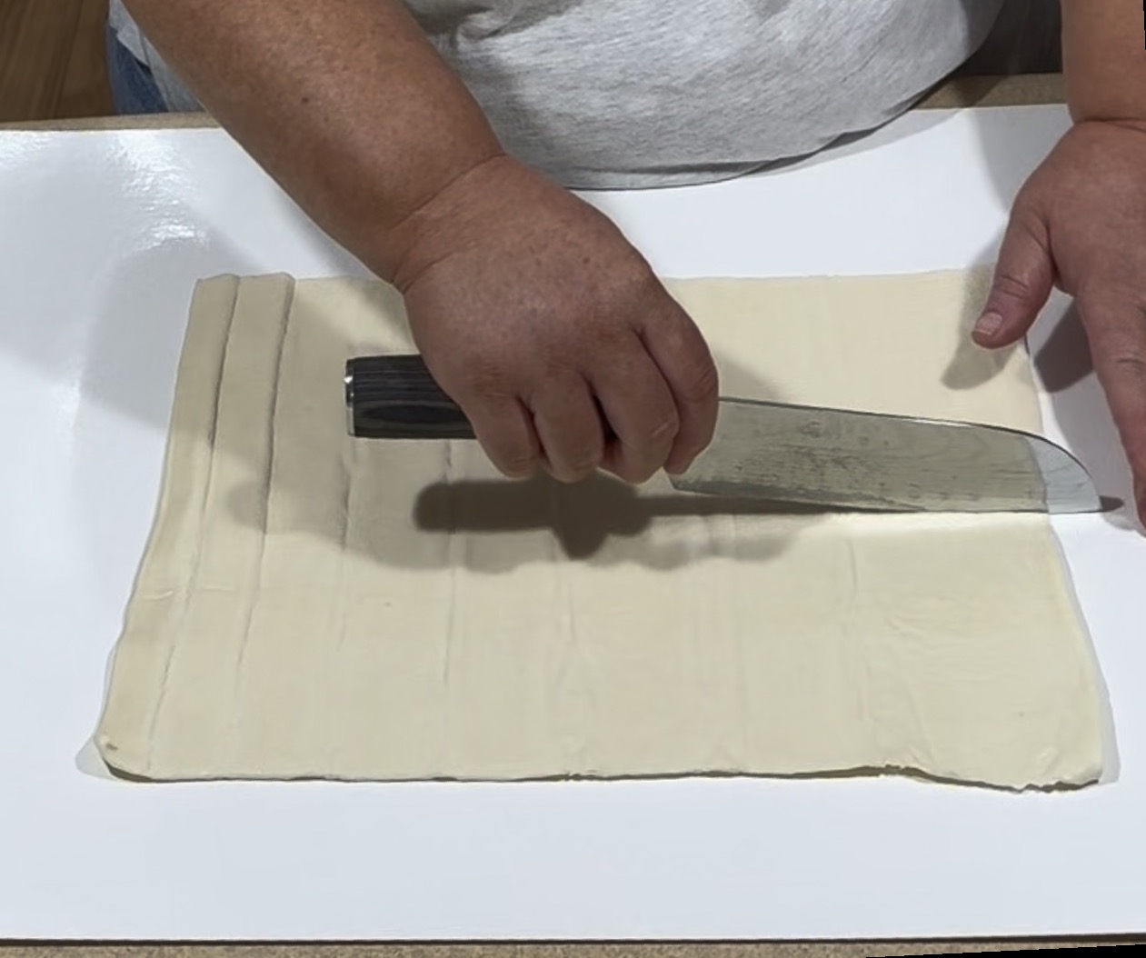 A person cuts rectangular strips into a sheet of raw dough on a white surface using a large kitchen knife.