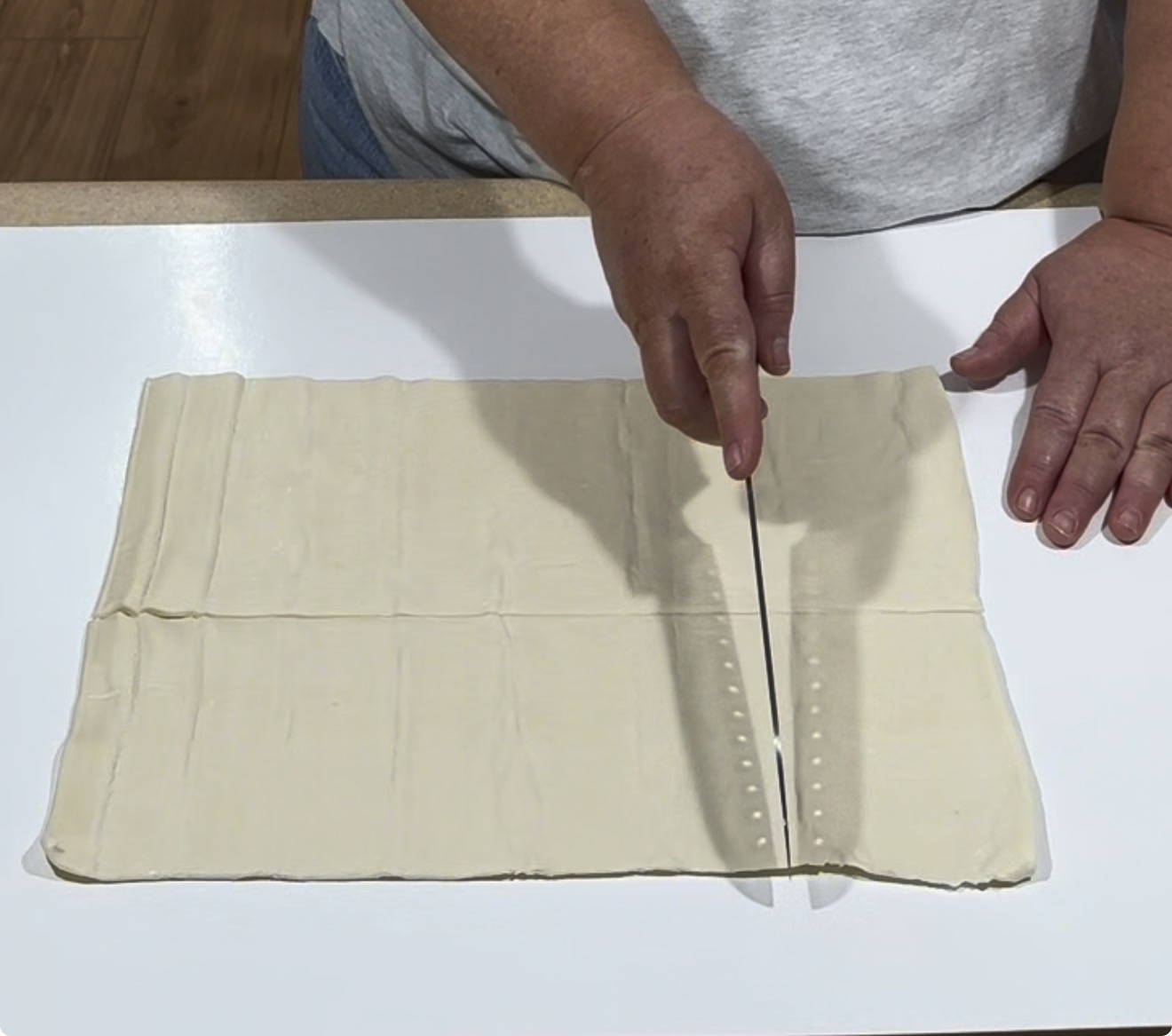 A persons hands cutting a rectangular sheet of puff pastry dough on a white surface with a large knife.