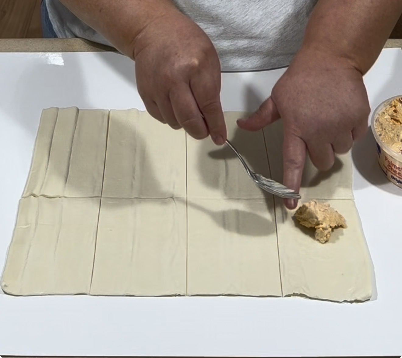 Hands placing a spoonful of filling onto a rectangular sheet of dough that has been cut into strips. The person is wearing a gray shirt and working on a white surface.