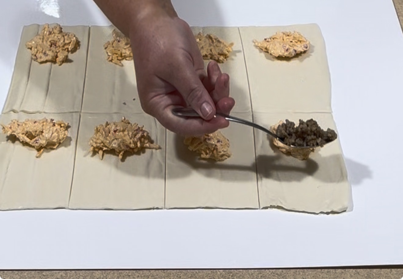 A hand uses a spoon to place a meat filling onto squares of dough, some already topped with a cheese mixture, on a white surface.