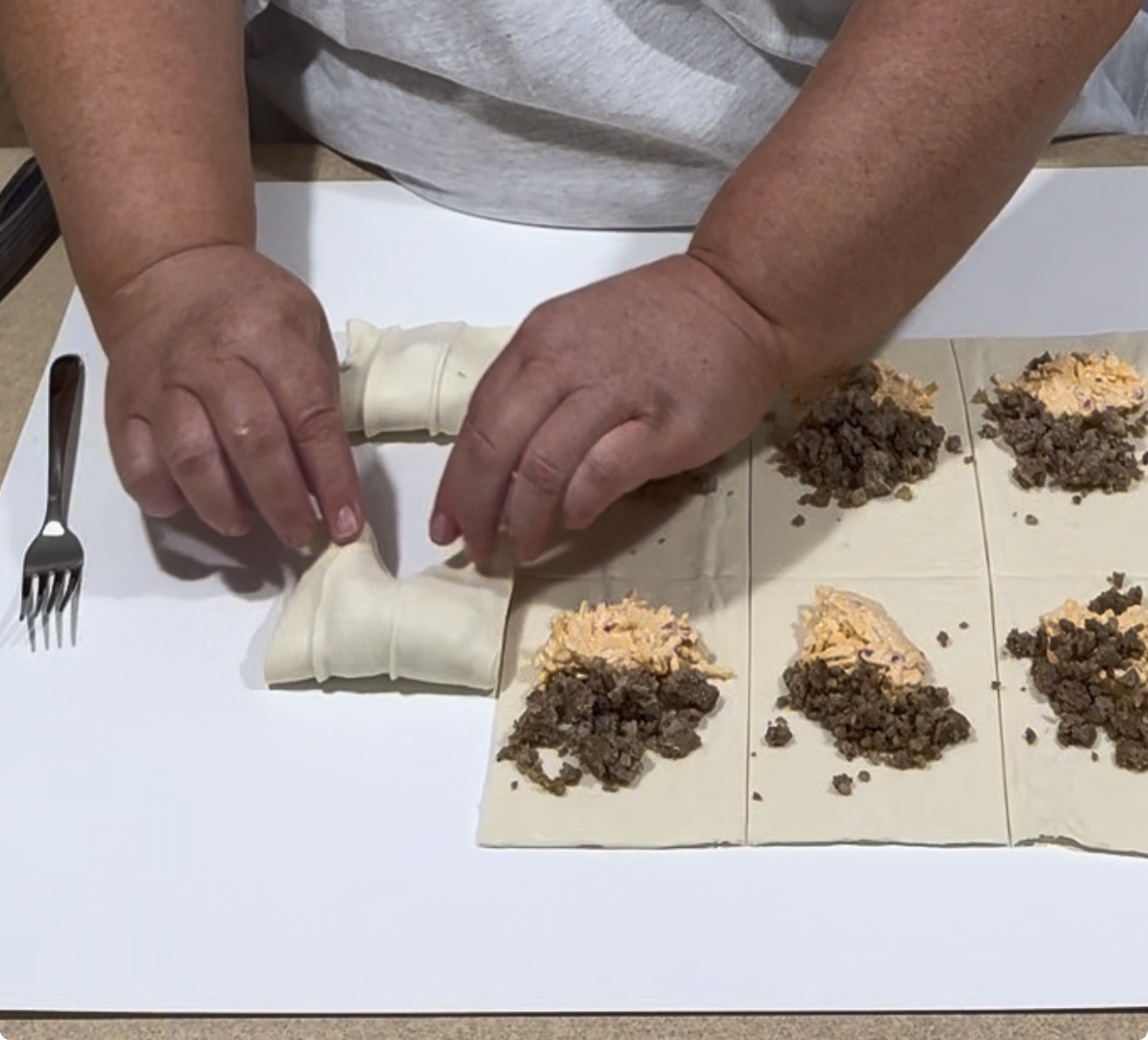 A person rolls dough filled with ground meat and cheese on a countertop, while four more portions of dough, meat, and cheese are prepared beside them. A fork is placed on the left.