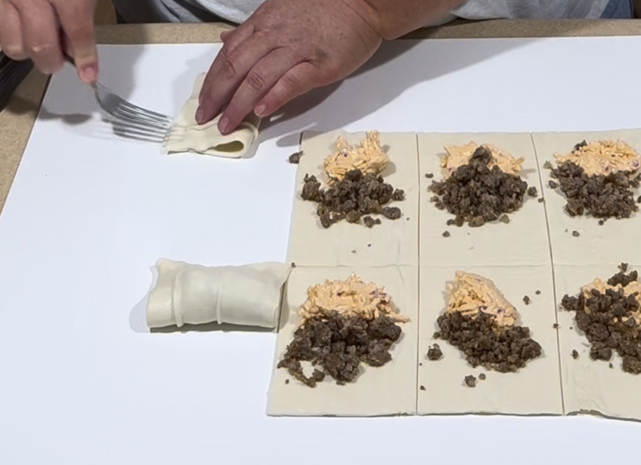 A person seals the edge of a dough square with a fork. Nearby, more dough squares are topped with cooked ground beef and orange shredded cheese on a white surface.