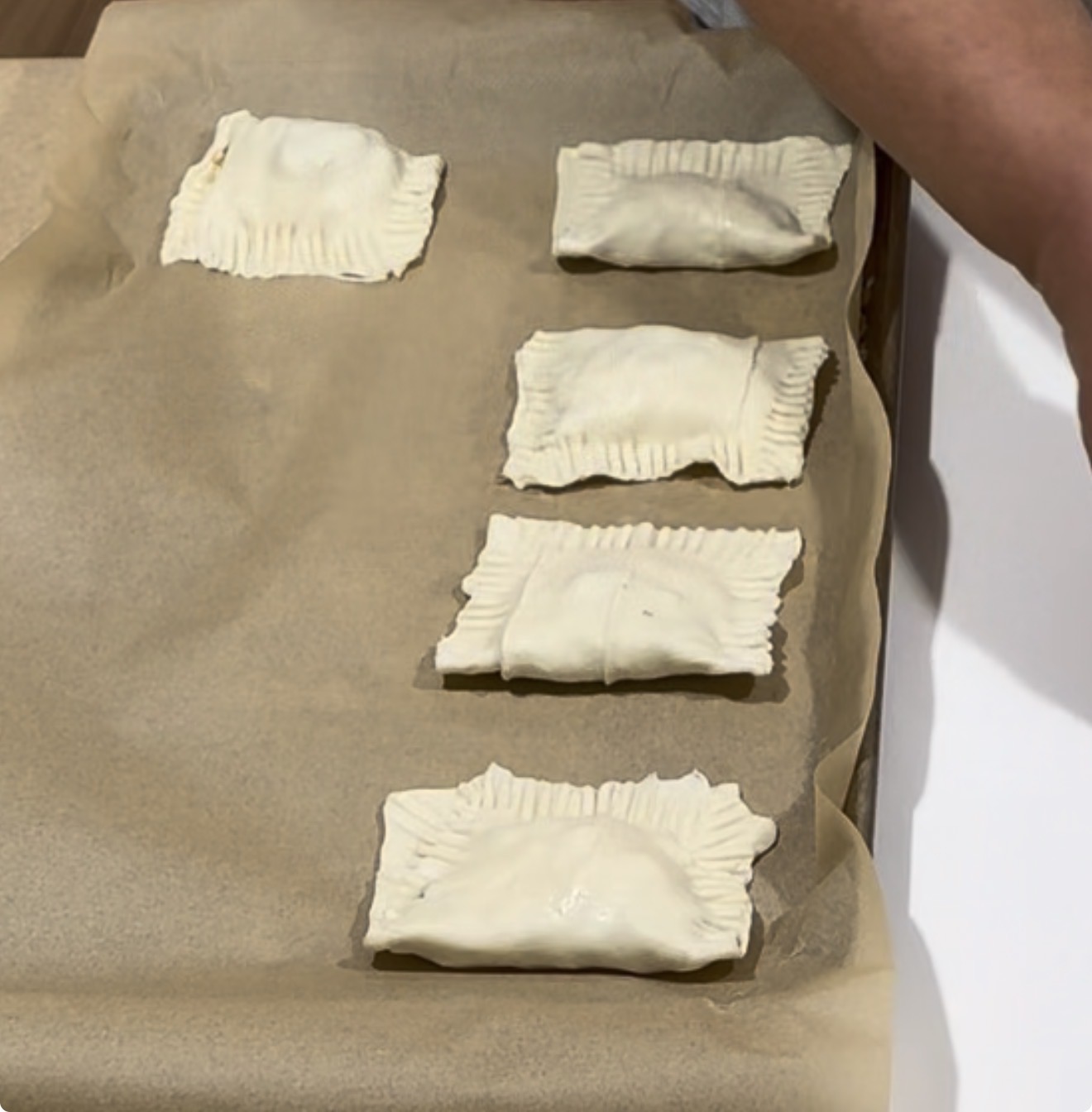 Four unbaked pastry rectangles on a parchment-lined baking sheet, each sealed with fork marks along the edges; a persons arm is visible on the right side of the image.