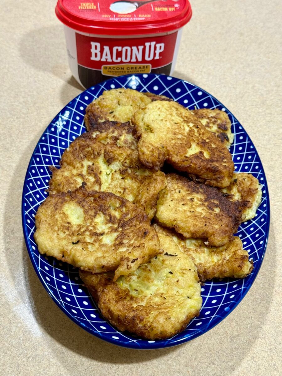 A blue patterned plate holds several golden-brown fritters. Behind the plate is a red tub labeled Bacon Up Bacon Grease on a beige countertop.