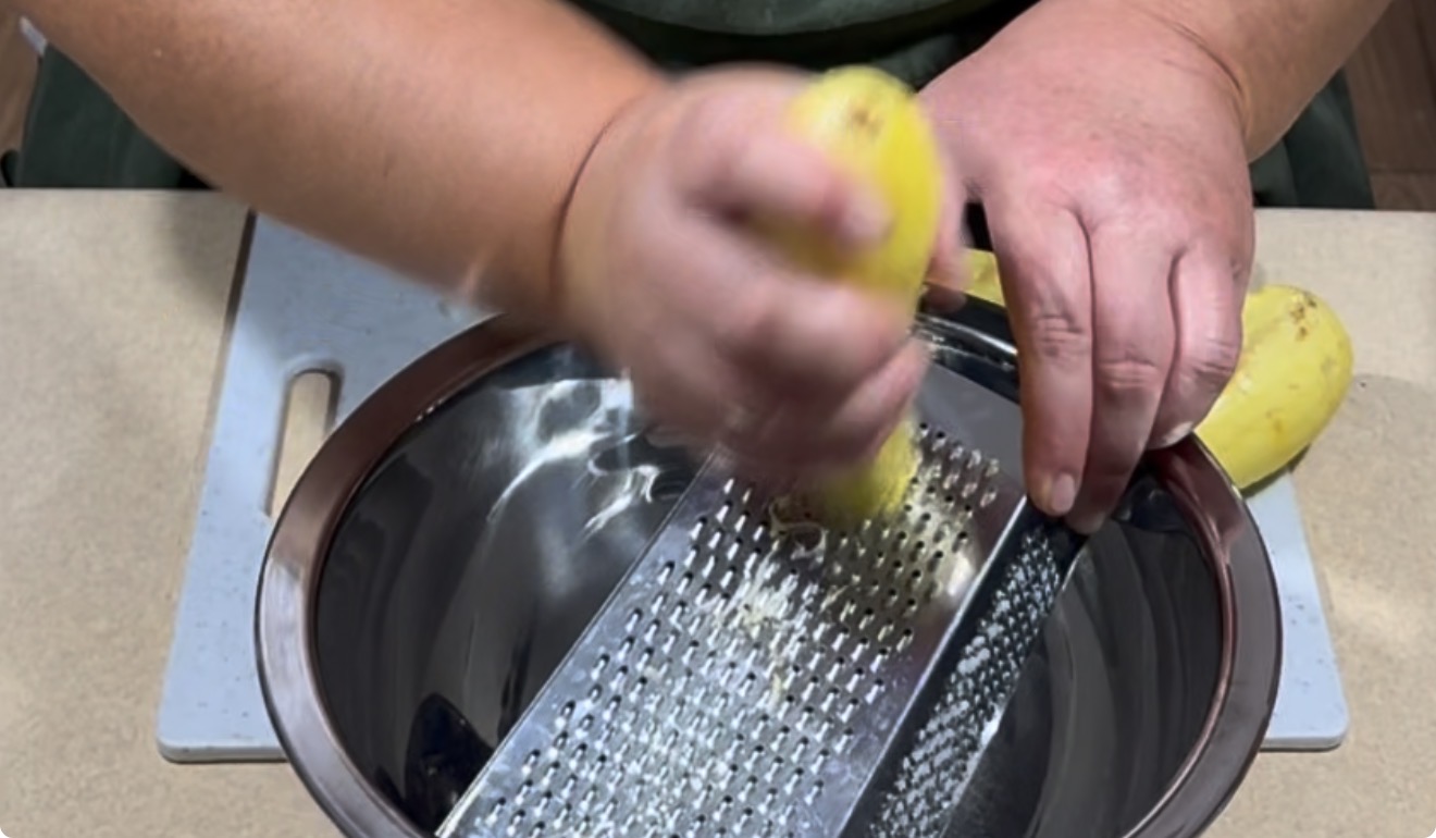 A person grates a yellow vegetable, possibly a potato, over a metal bowl using a box grater, with a cutting board underneath.