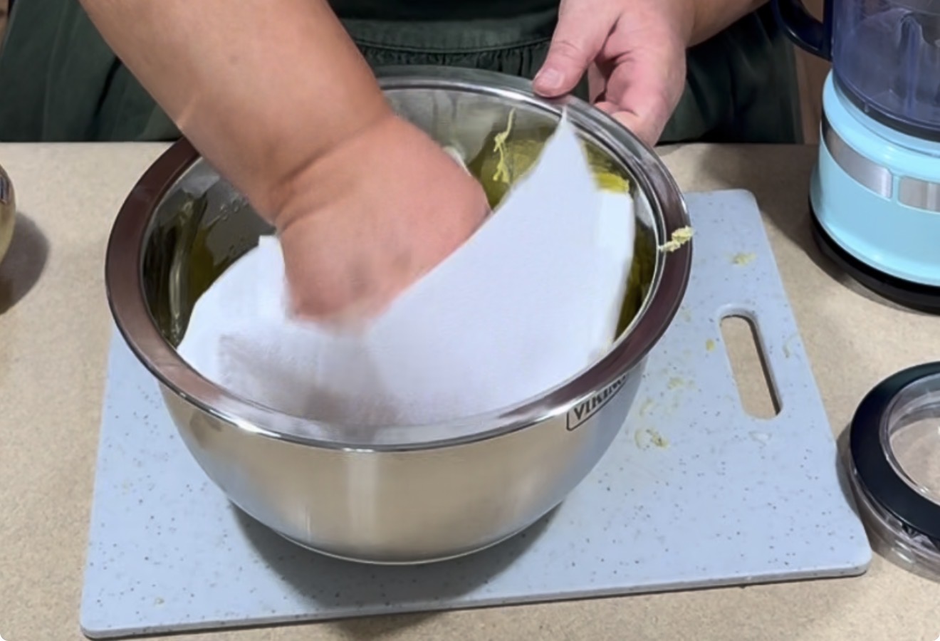 A person wipes the inside of a stainless steel mixing bowl with a white paper towel on a light-colored cutting board, next to a blue kitchen appliance.