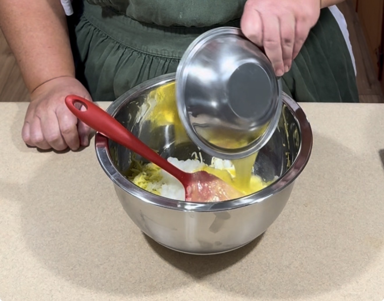 A person in a green apron pours beaten eggs from a metal bowl into a larger mixing bowl with other ingredients, using a red spatula to mix on a beige countertop.