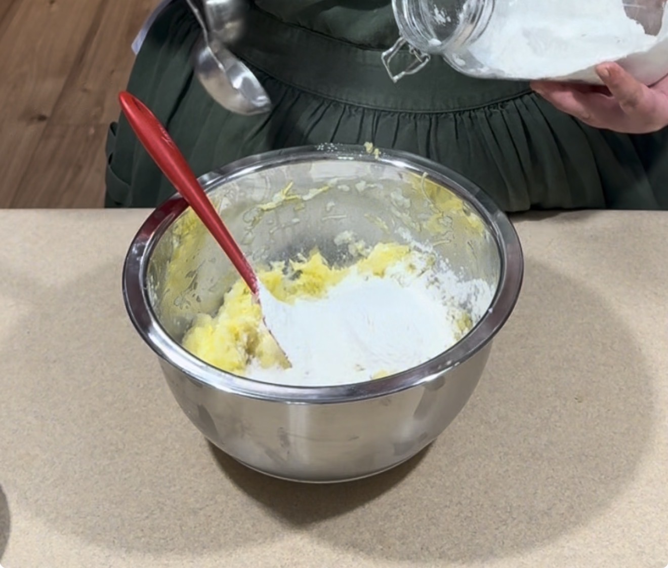 A person adds flour from a glass jar into a metal mixing bowl with a red spatula and yellow batter, preparing ingredients on a kitchen counter.