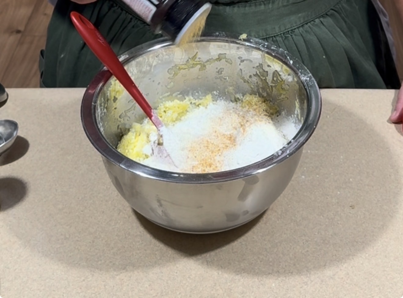 A person pours grated cheese from a container into a mixing bowl filled with a yellow mixture, holding a red spatula. The bowl rests on a countertop.