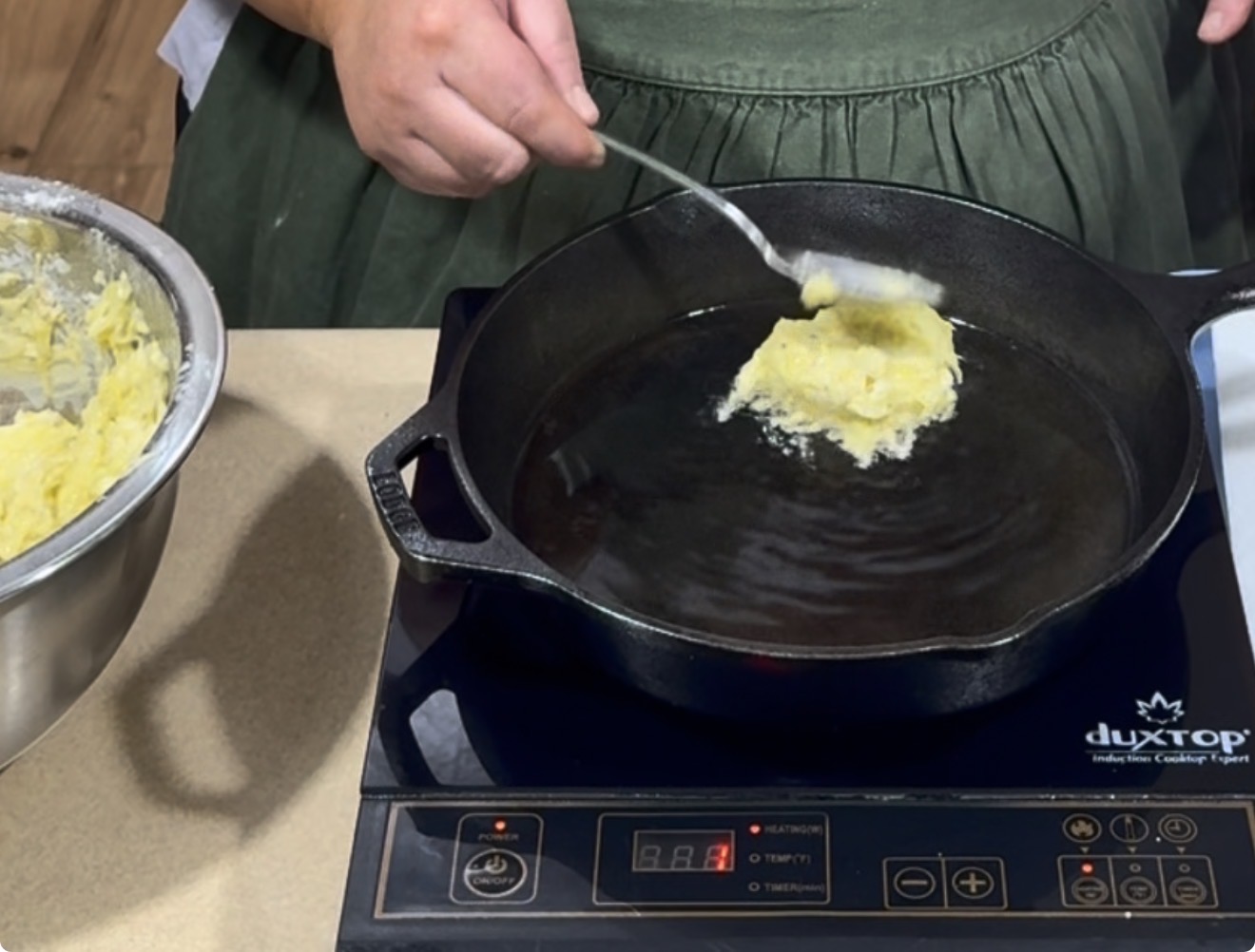 A person spoons batter from a bowl into a cast iron skillet with oil on an induction cooktop, preparing to fry the mixture.