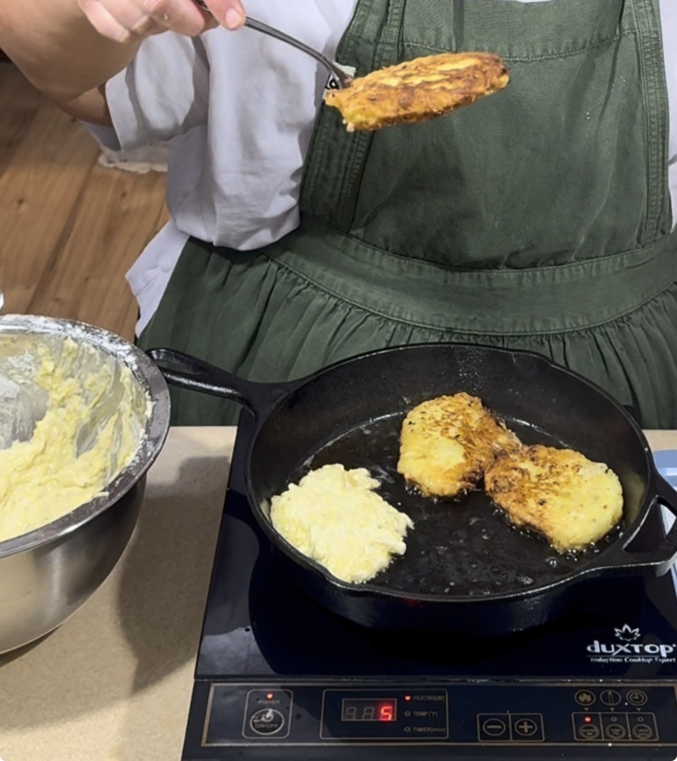 A person in a green apron fries potato pancakes in a black skillet on an induction stove, with a bowl of batter beside them.