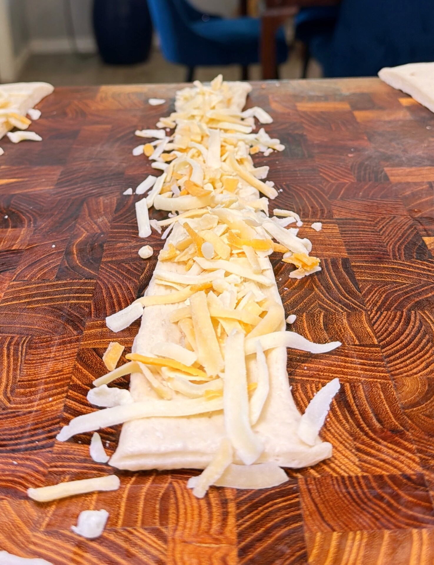 Shredded cheese scattered on a strip of dough placed on a wooden cutting board, ready to be rolled or baked.