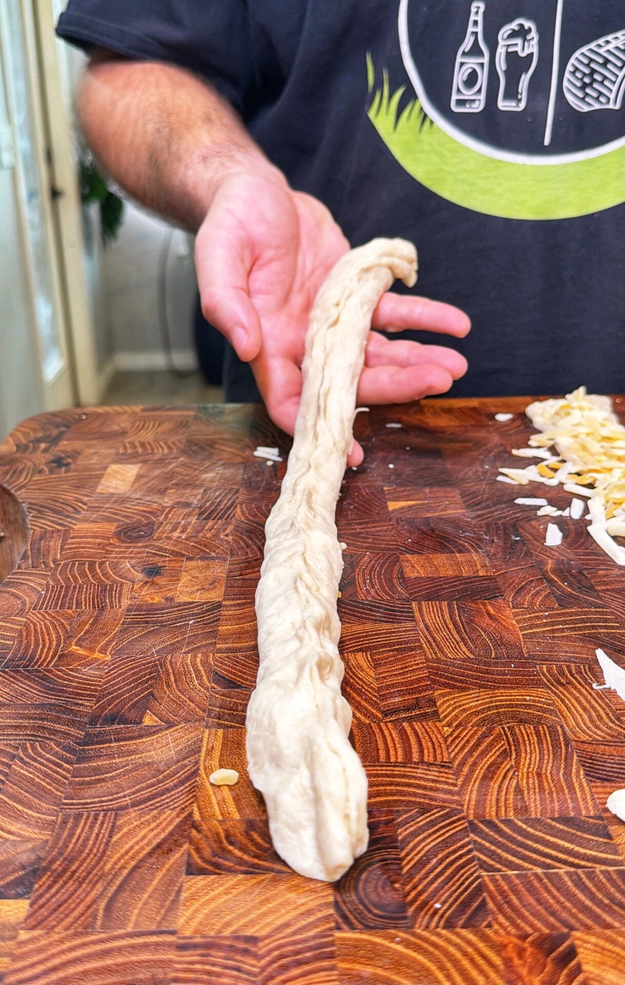 A person holds a long, twisted piece of dough over a wooden cutting board scattered with grated cheese.
